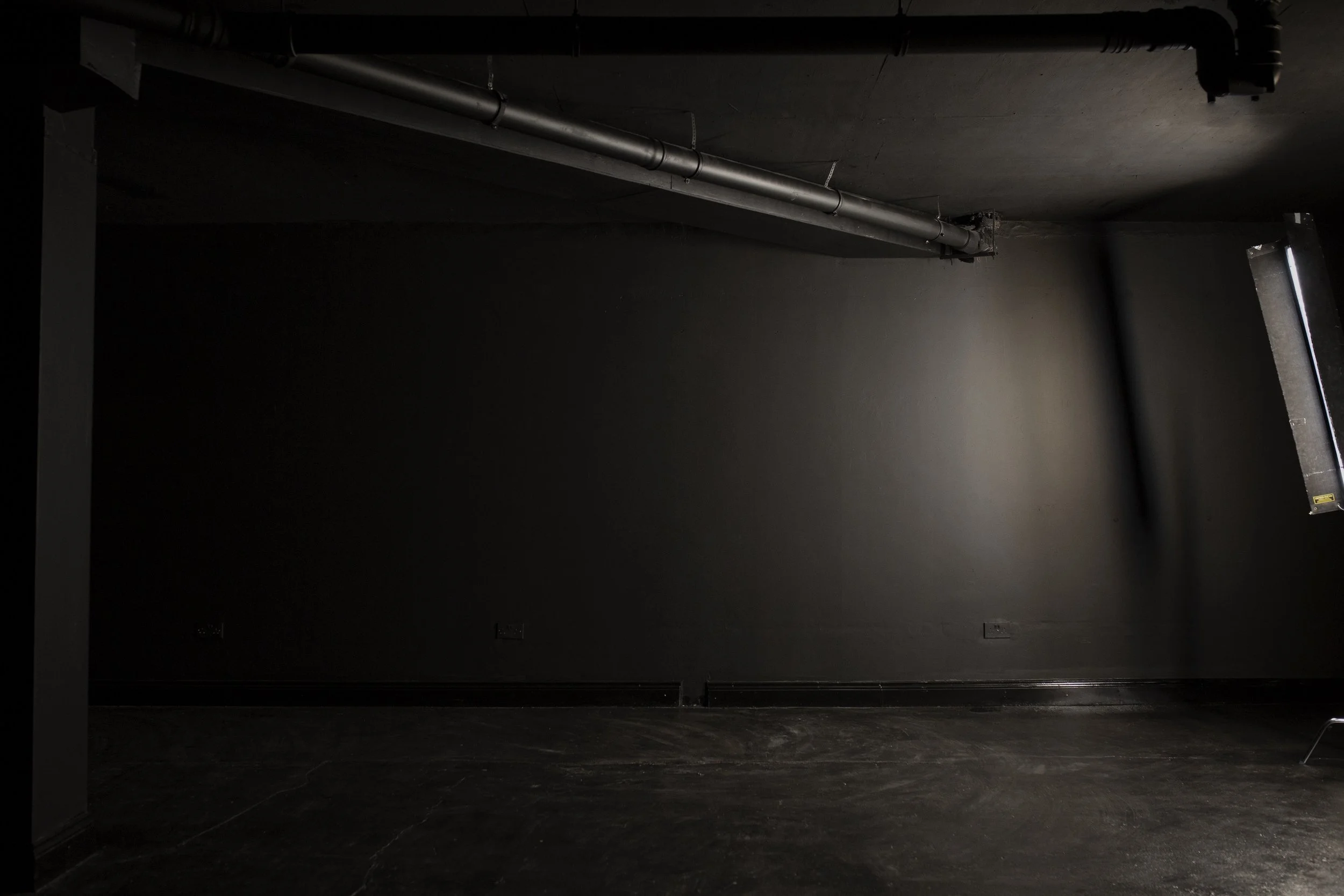 Empty dark room with a black wall, exposed pipes on the ceiling, and a partially open window on the right.