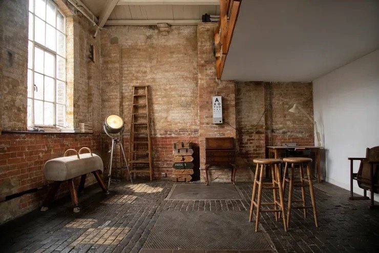 A rustic industrial-style interior with exposed brick walls, a ladder, a spotlight, a wooden table with two stools, and a framed eye chart.