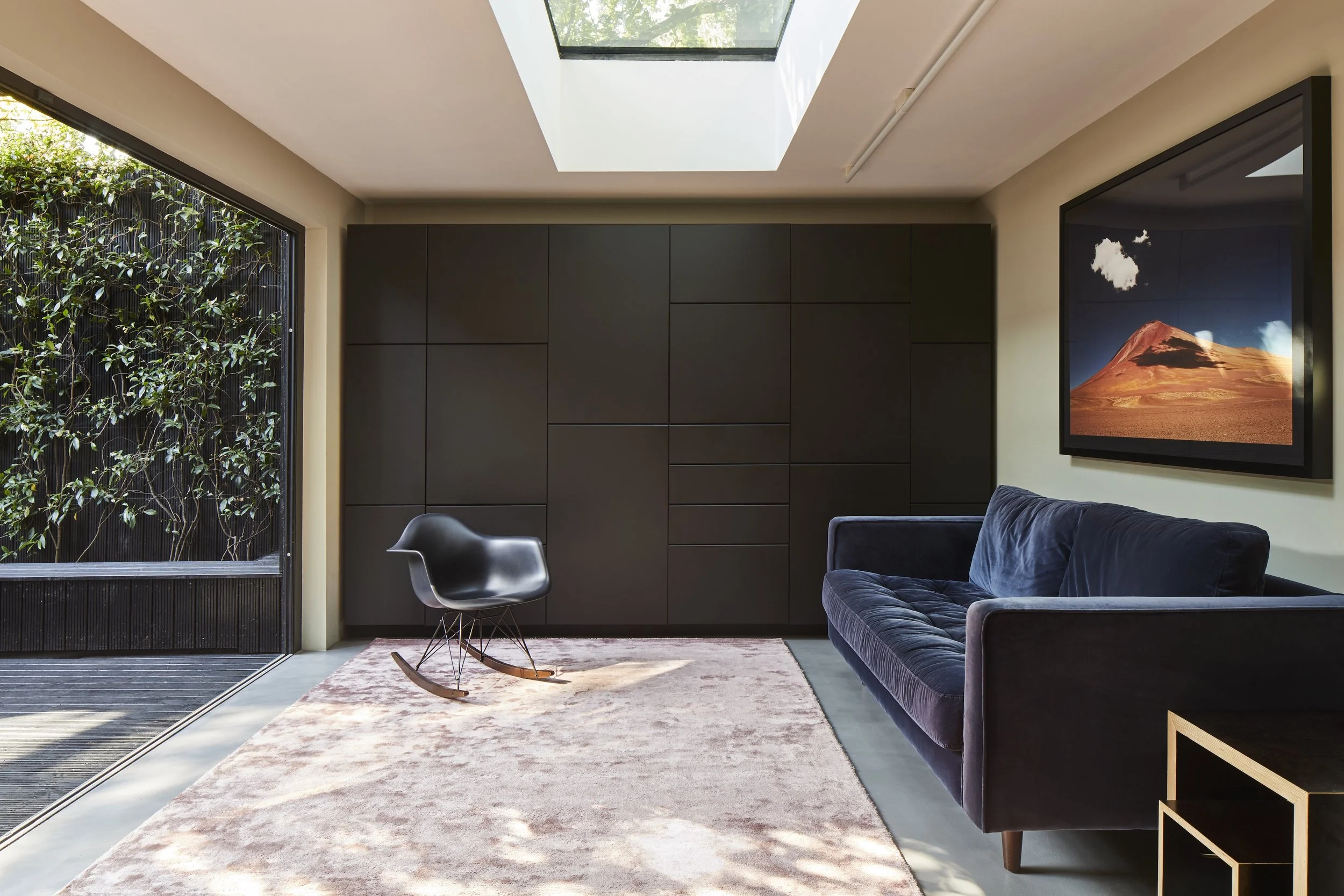Modern living room with a large window, black armchair, navy velvet sofa, artwork of a mountain on the wall, and a skylight.