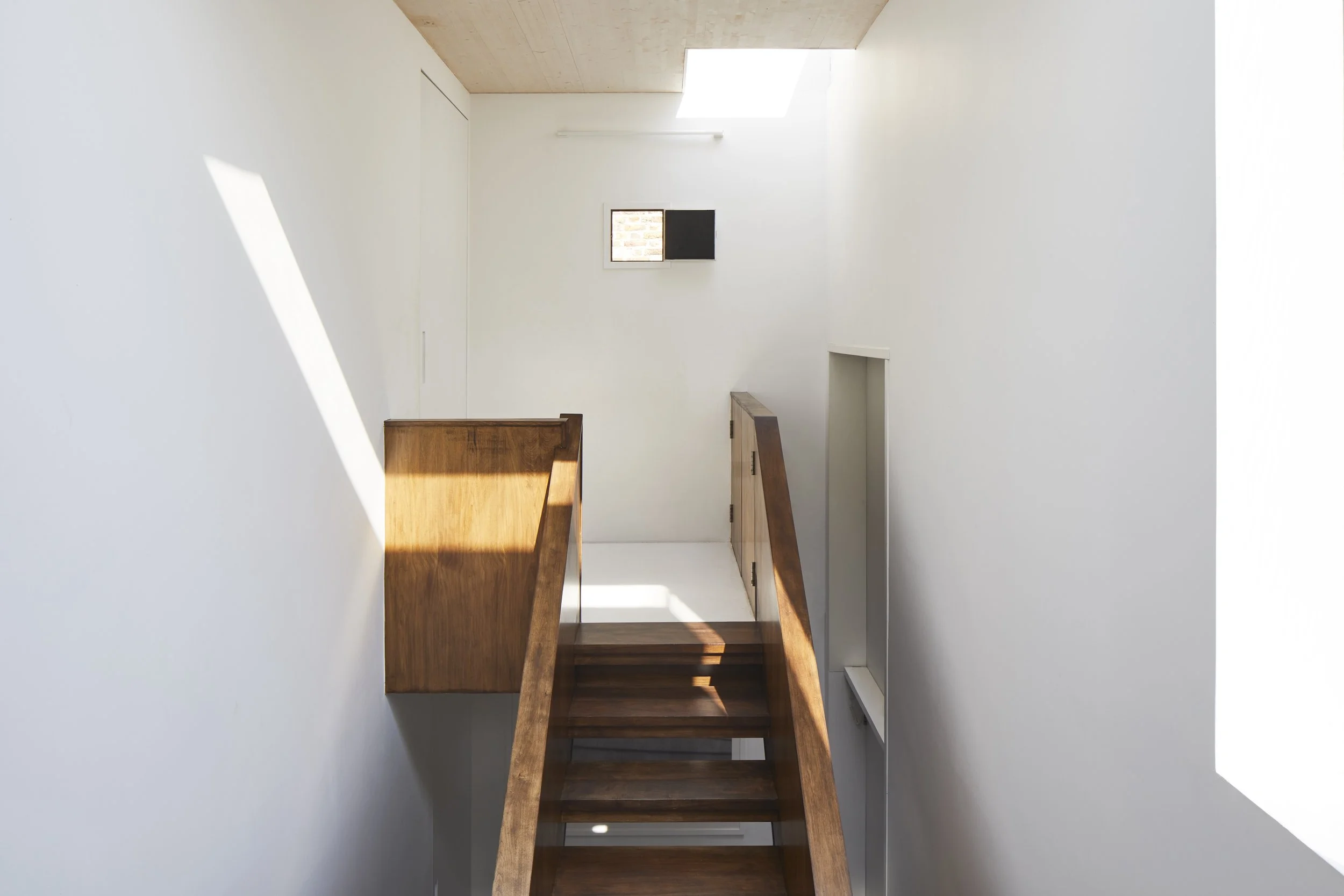 Interior view of a staircase with wooden handrails and steps, white walls, a small window, and skylights allowing natural light into the space.