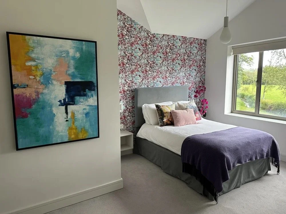 A bedroom with a large window showing green trees and grass outside. The bed has a gray headboard, white sheets, and decorative pillows, with a purple throw blanket at the foot. The wall behind the bed features floral wallpaper in shades of pink, red