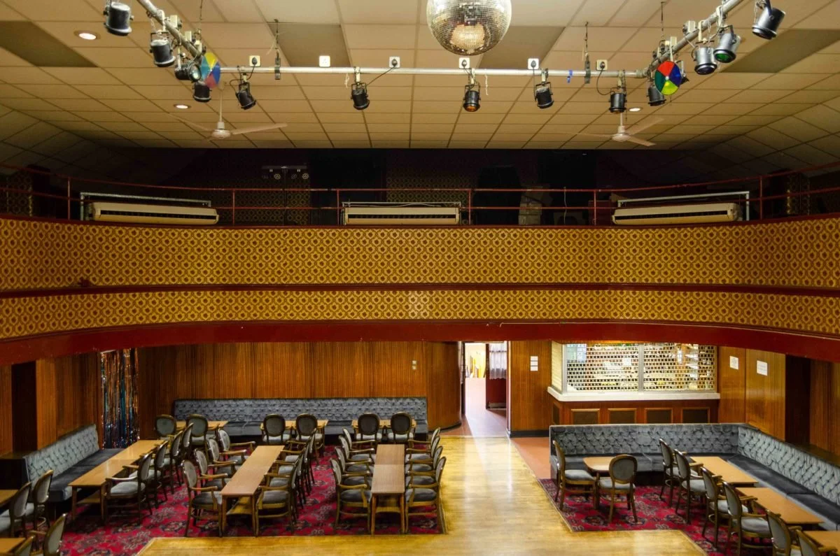 An empty nightclub dance floor with a band stage, a lower level with tables and chairs, and a balcony with additional seating, decorated with wood paneling, patterned wallpaper, and a disco ball hanging from the ceiling.
