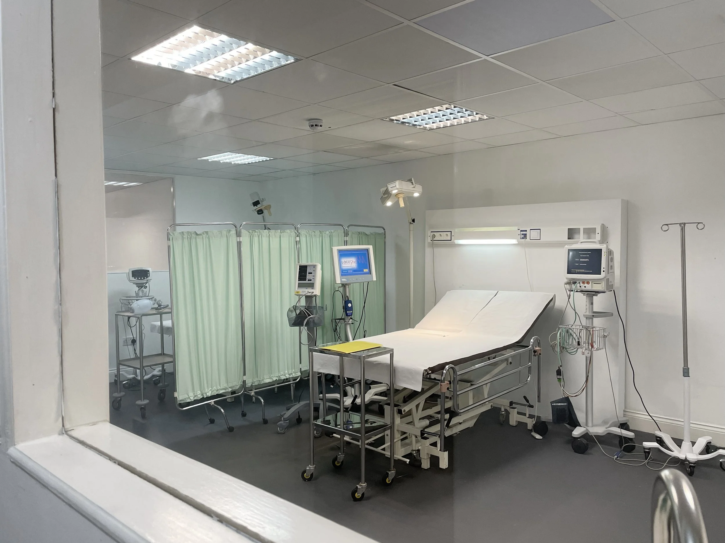 Hospital room with medical equipment, hospital bed, monitors, IV stand, and privacy curtains, seen through a window.