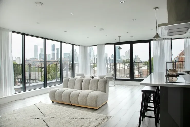 Modern living room with large windows showing city skyline, beige sofa, kitchen counter with stools, and white curtains.