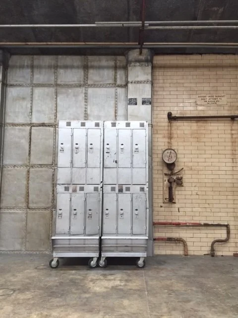A set of storage lockers on wheels leans against a concrete wall in an industrial setting, with pipes and a gauge on a brick wall to the right.
