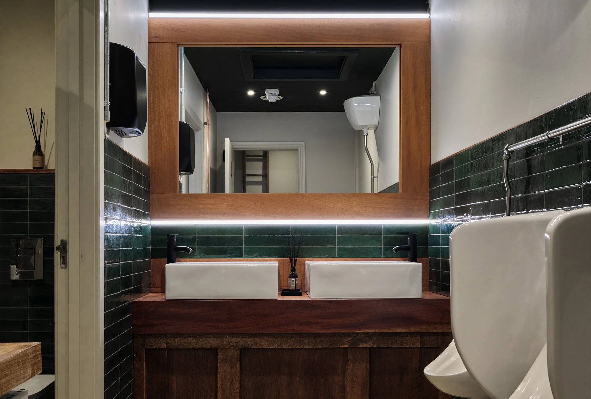 Public restroom with twin sinks, large mirror, green tiled walls, white urinal, and hand dryer, with a wooden frame around the mirror.
