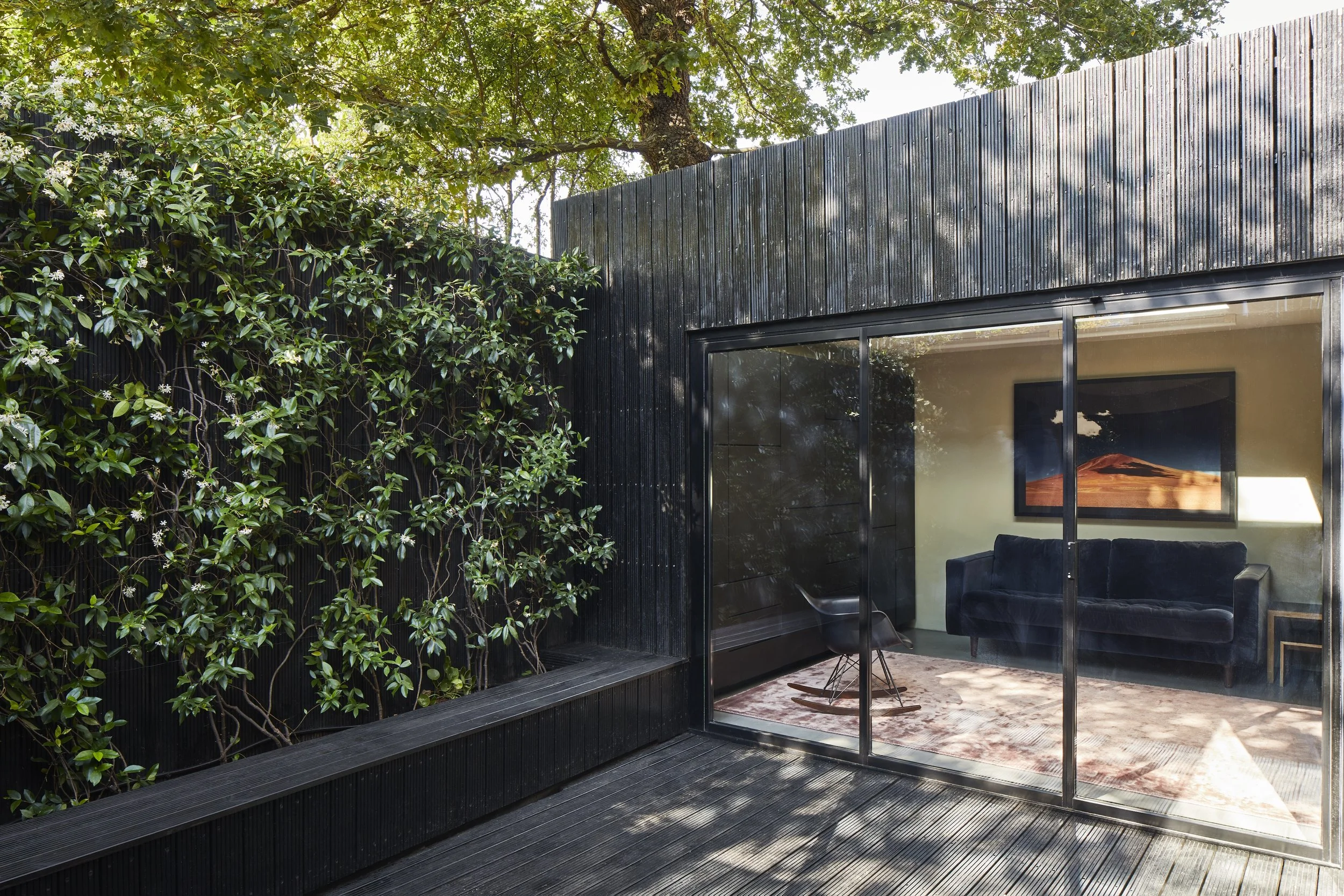 Exterior view of a modern house with black siding, large glass sliding doors, a small wooden deck, and a green garden wall with trees and vines.