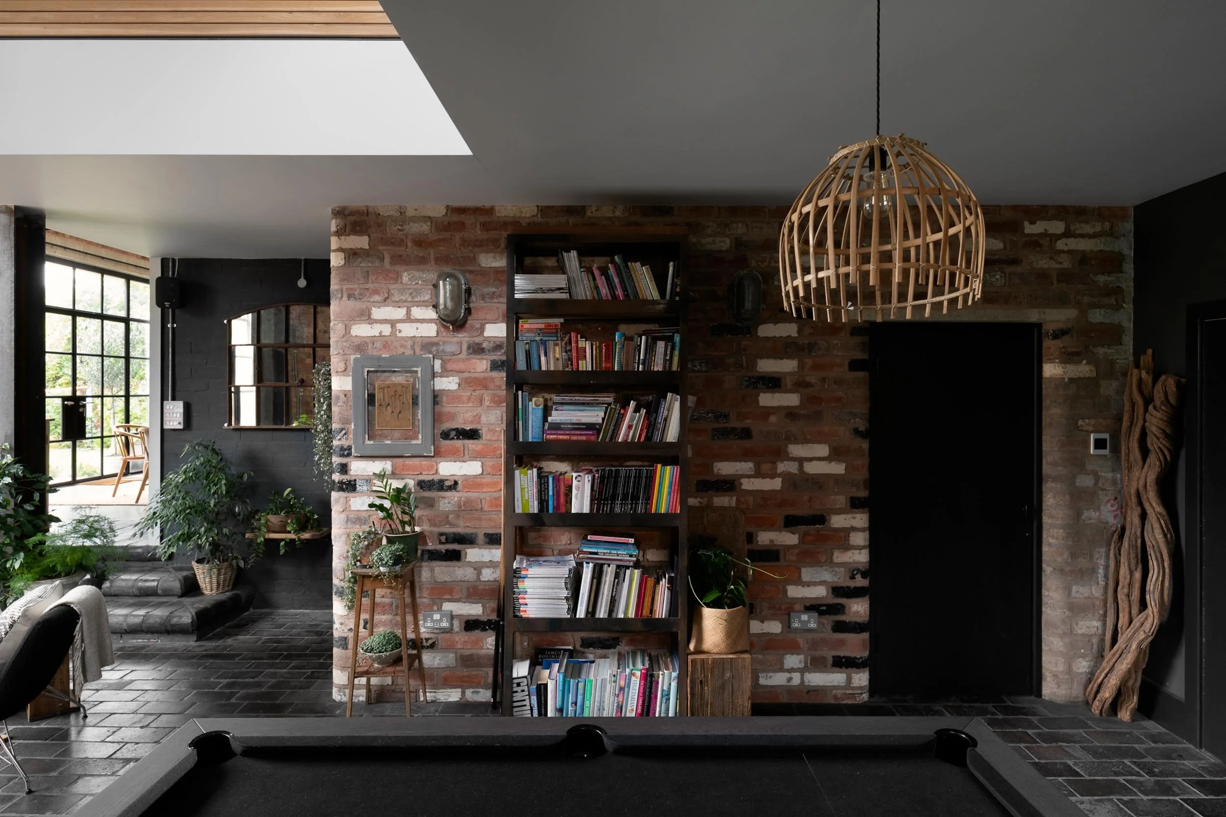 Interior living space with exposed brick wall, black door, bookshelf filled with books, plants, wooden decor, black pool table in foreground, large window with outdoor seating area, and ceiling light fixture.
