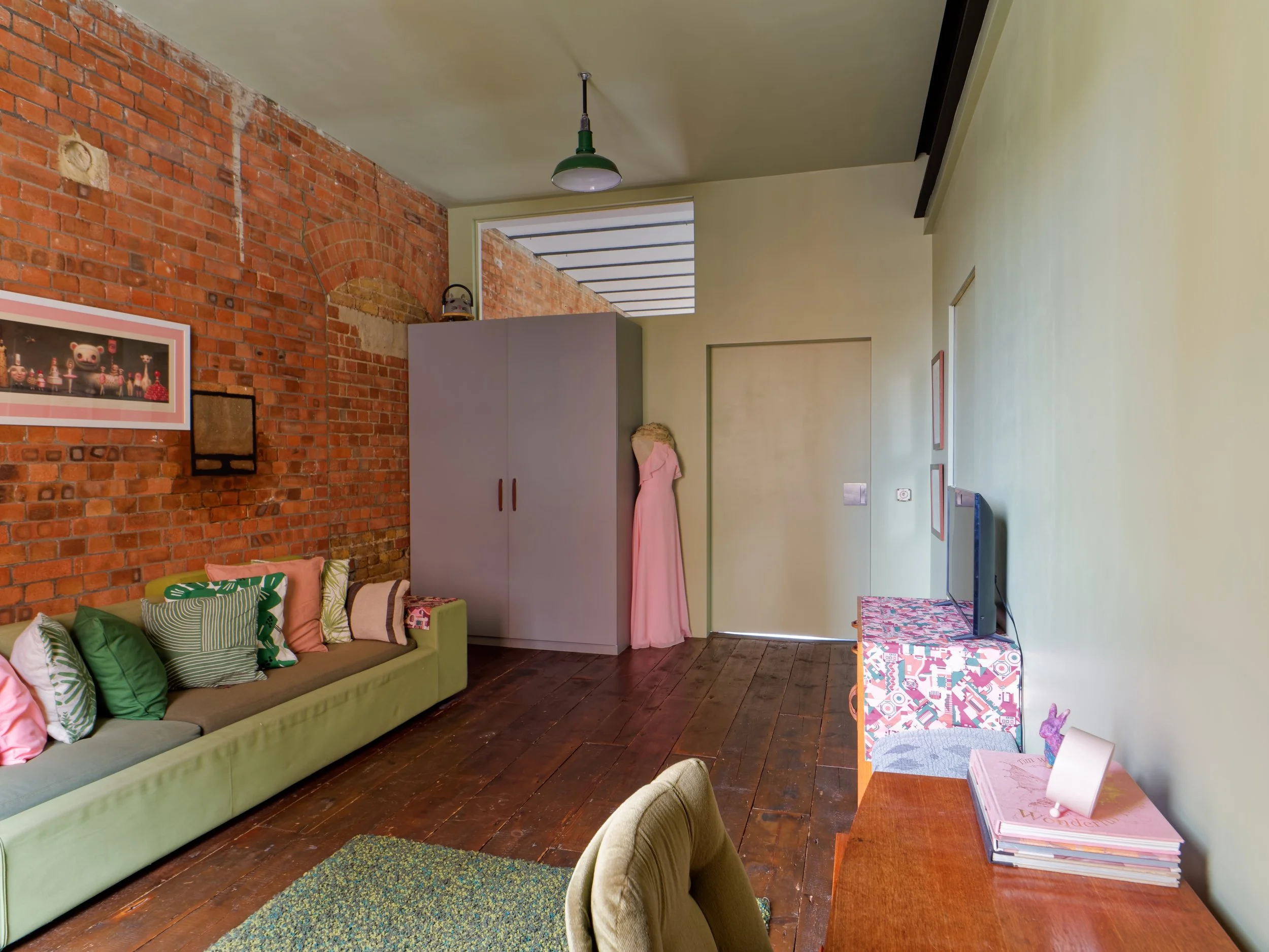 A room with a brick accent wall, a long light green sofa with assorted pillows, a wooden table with a stack of books and framed pictures, a small TV on a colorful patterned table, a closed door, and a pink dress hanging on a wardrobe. There is a ceil