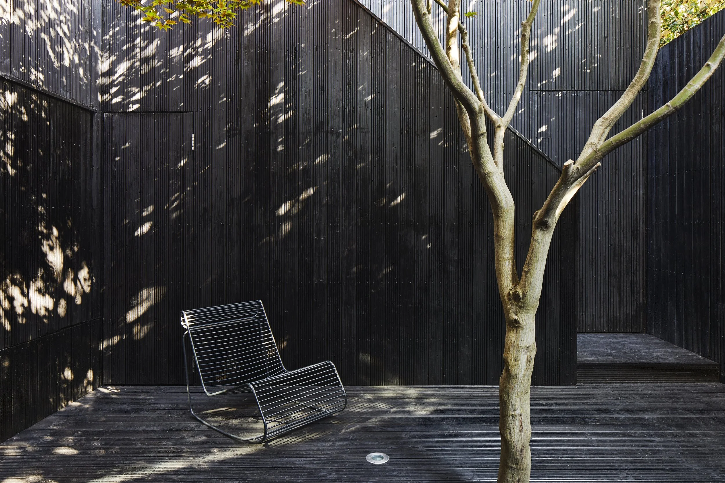 Minimalist outdoor patio with black wooden walls, a metal lounge chair, a tree, and sunlight casting shadows.