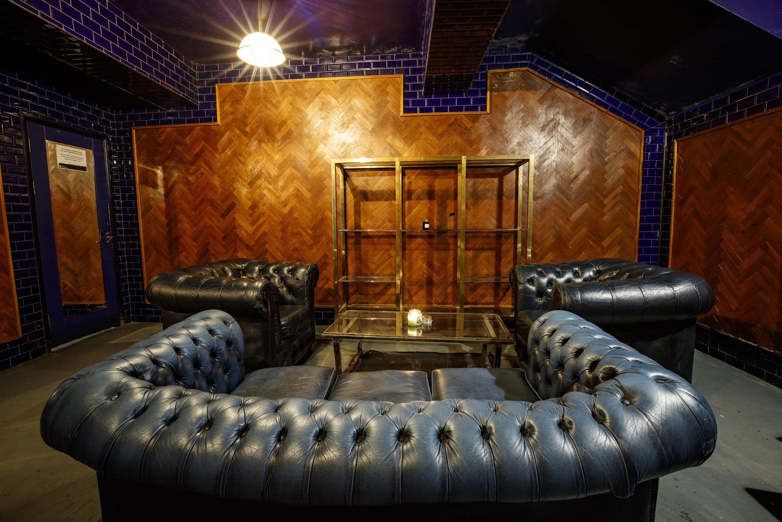 A cozy lounge area with four black leather tufted sofas arranged around a glass coffee table in front of a wooden-paneled wall with blue tiled accents and a shelf, lit by a ceiling light.