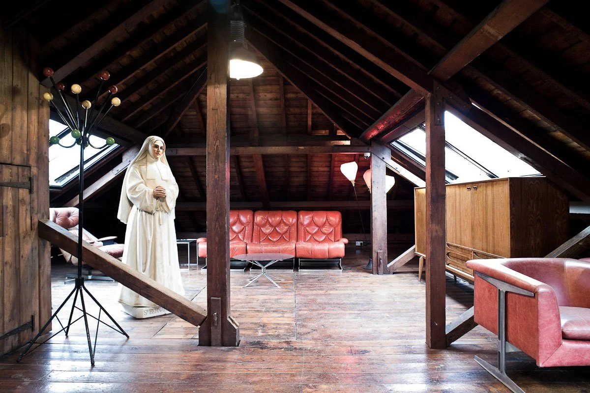 An attic room with wooden walls and ceiling, illuminated by two skylights and a hanging light fixture. The room features a vintage-style mannequin dressed as a nun, a pink upholstered sofa, a pink armchair, a small wooden cabinet, a coat rack, and va