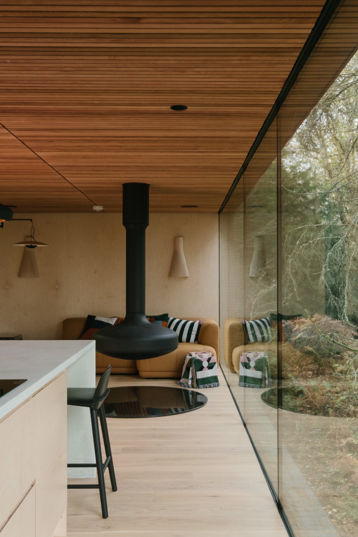 Modern living room with large glass wall showing trees outside, wooden ceiling, beige walls, yellow sofa with striped cushions, pink throw blanket, wall-mounted light fixtures, and black hanging fireplace