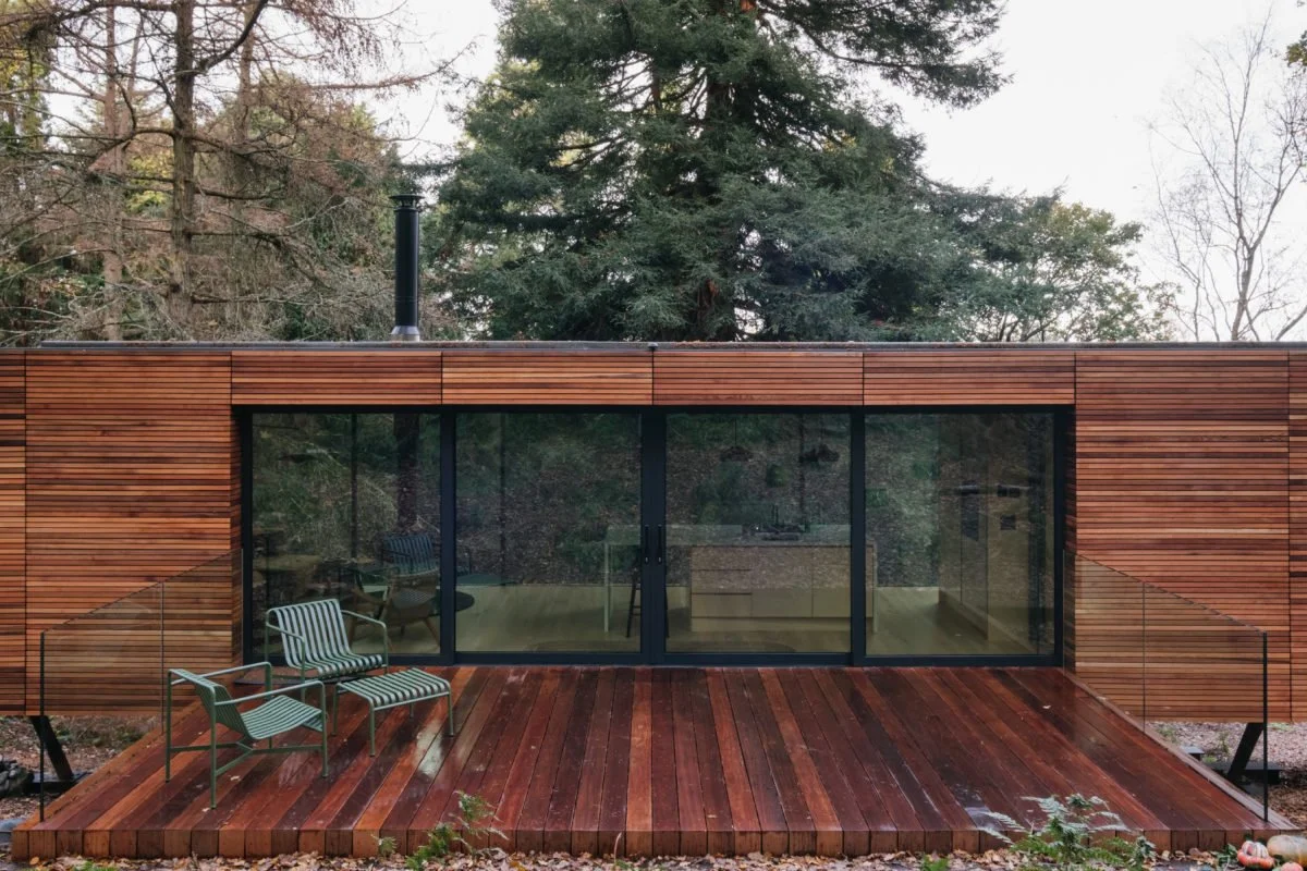 Modern house with large glass sliding doors, wooden exterior, and a wooden deck outdoor seating area with two chairs, surrounded by trees.