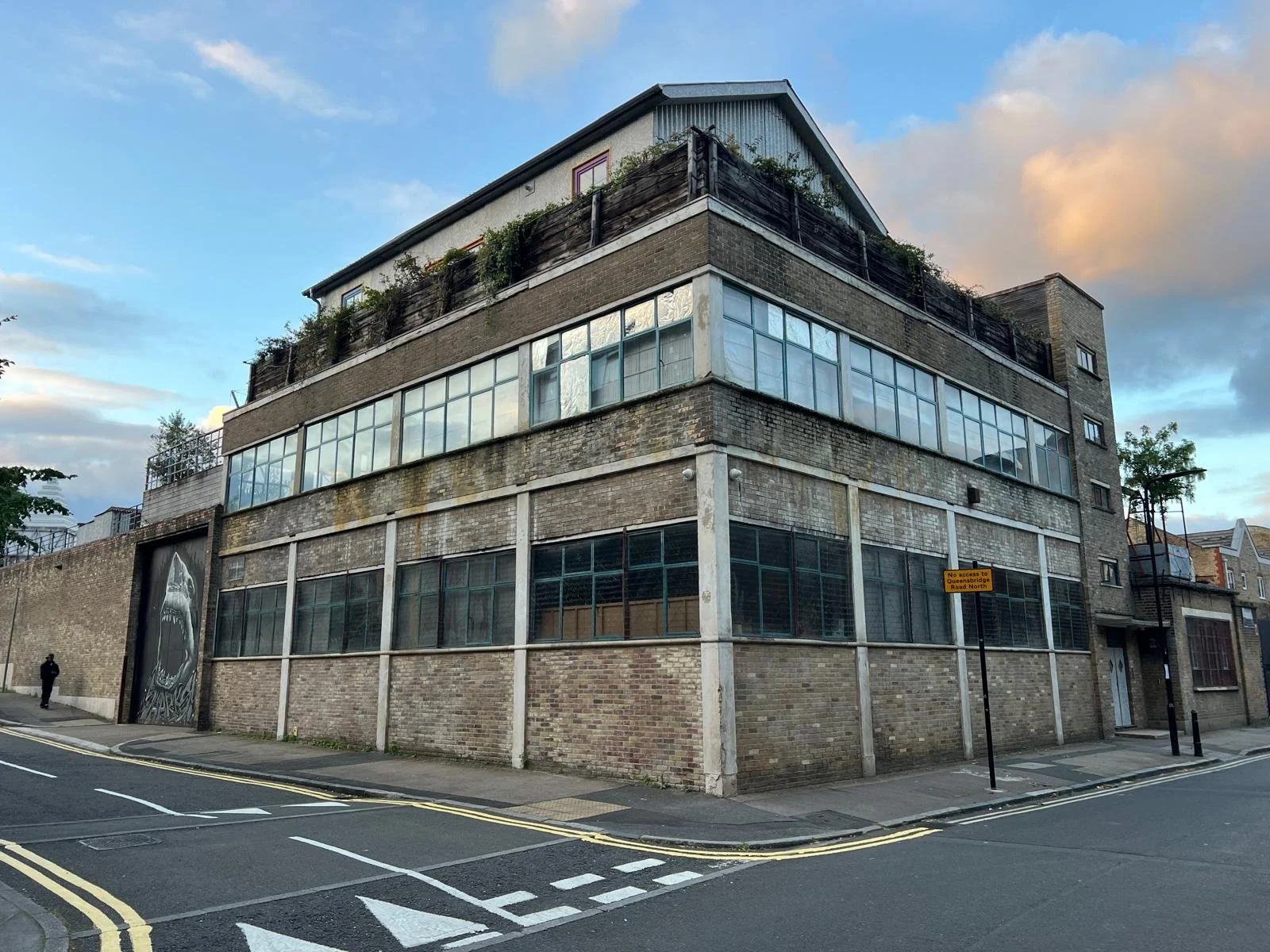 A multi-story brick building with large windows on each floor, situated at the corner of a street with yellow road markings. A person is walking on the sidewalk near the building. The sky is partly cloudy with some sunset lighting.