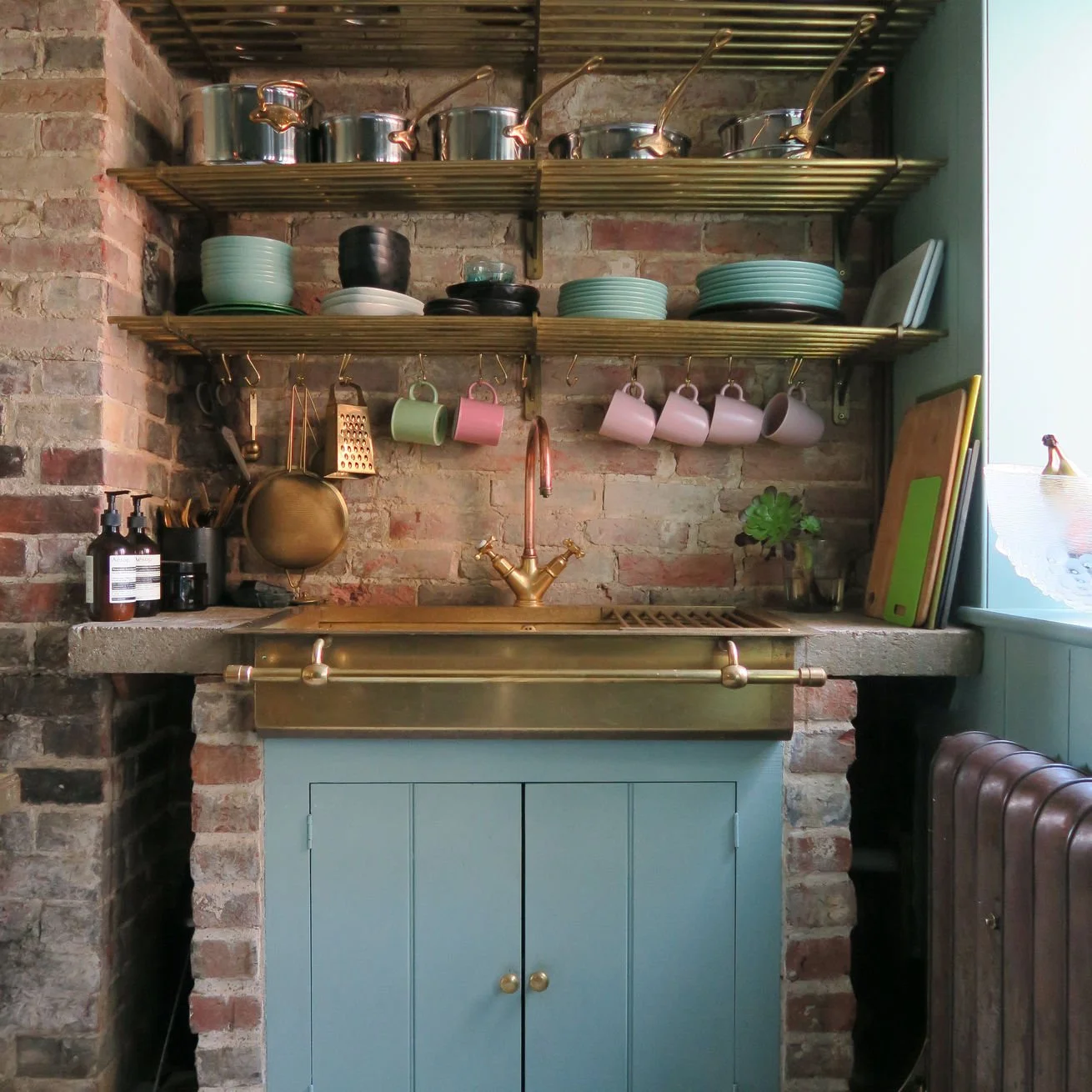 A cozy kitchen corner with brick wall, blue cabinet, brass sink, hanging colorful cups, plates, and kitchen utensils, and open shelving with cookware.