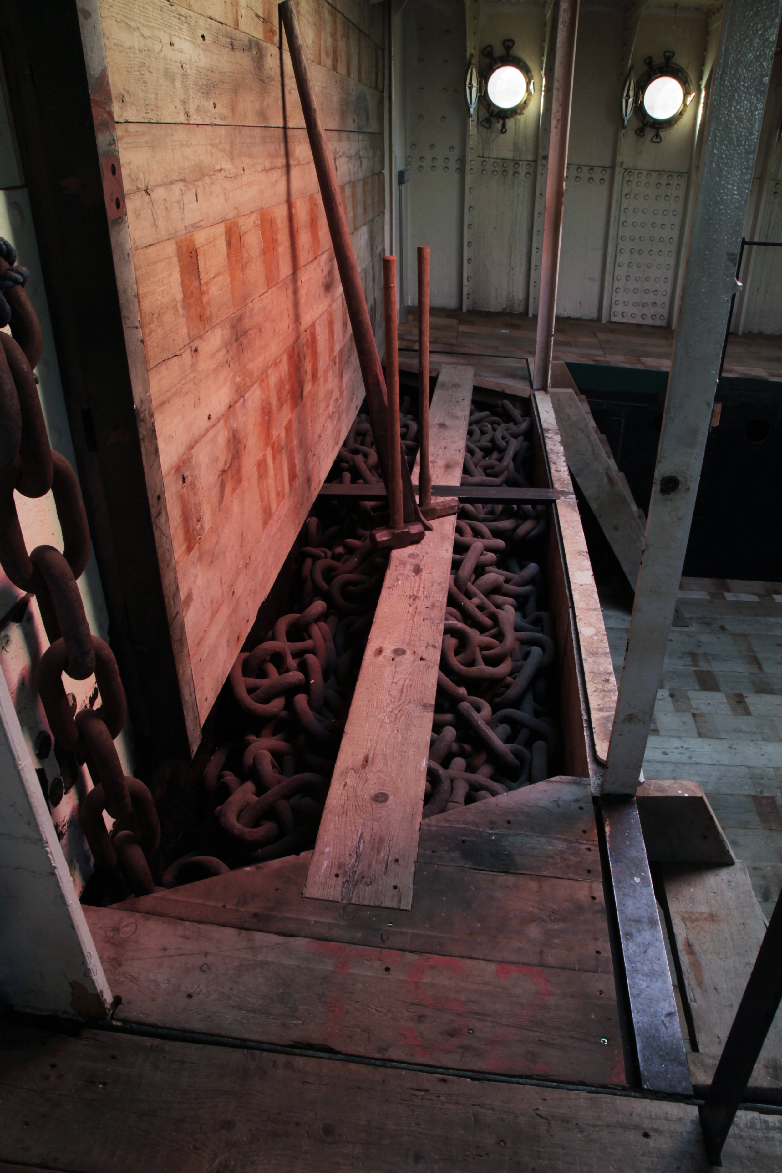 Interior of a ship showing a large pile of heavy chains, with some wooden boards and metal structures around.
