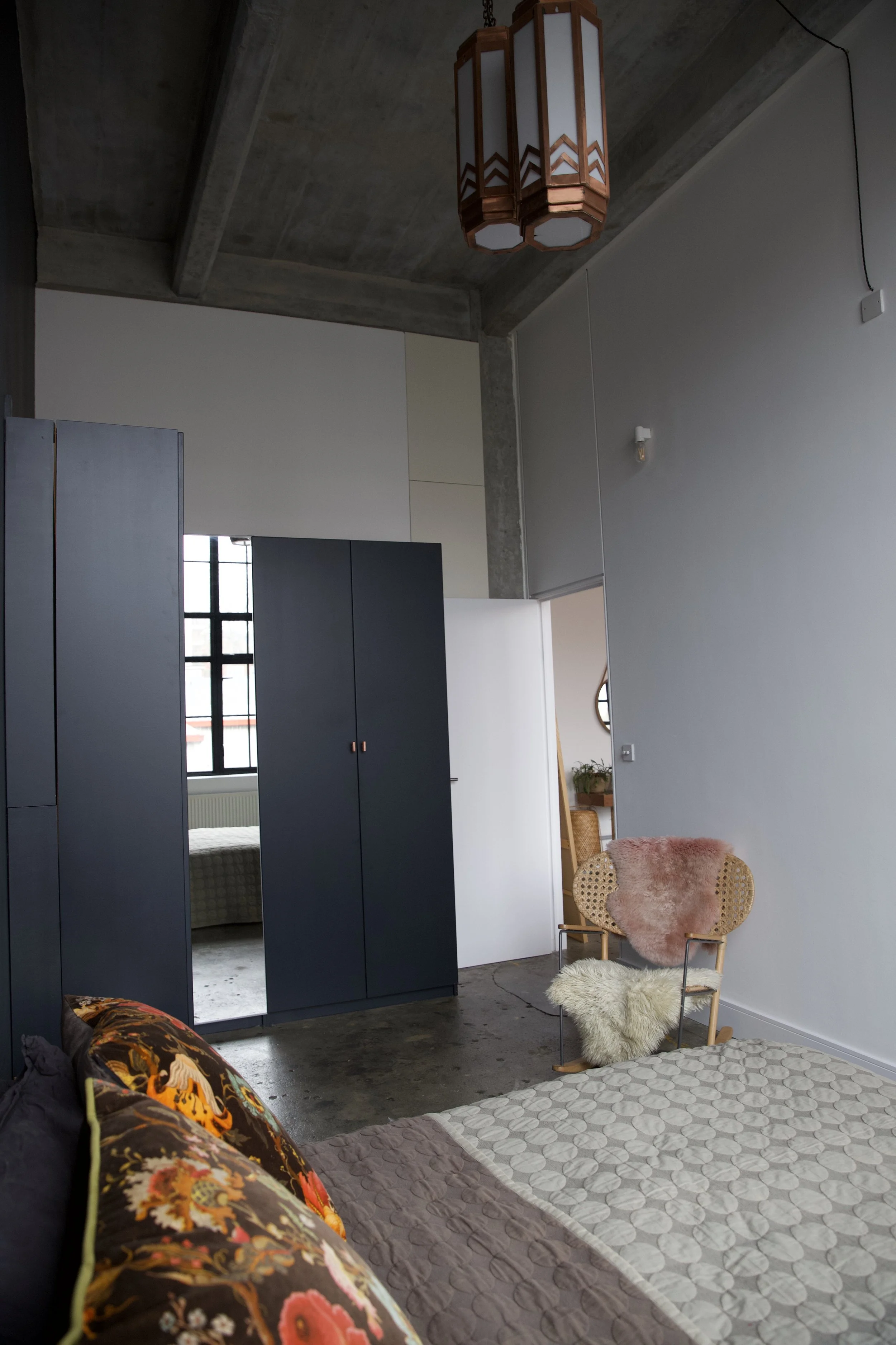 Interior of a minimalistic bedroom with a bed, colorful pillows, a rocking chair with plush throws, dark wardrobe, and modern light fixture on a concrete ceiling.