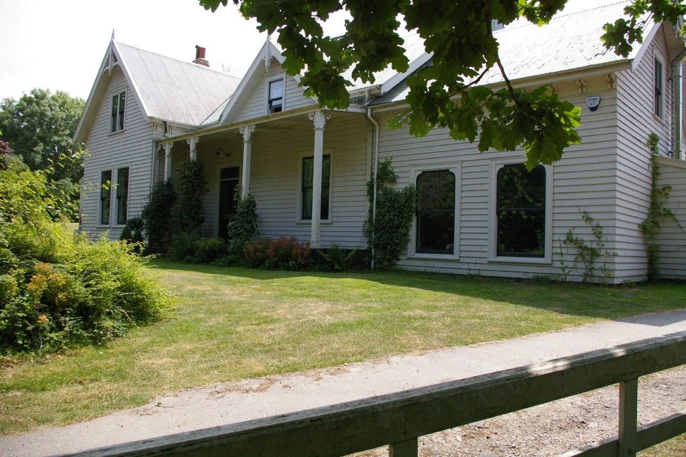 A large white Victorian-style house with bay windows, a metal roof, and decorative trim. The house has a front porch supported by columns, surrounded by green shrubbery, trees, and a lawn, with a sidewalk or driveway in front.