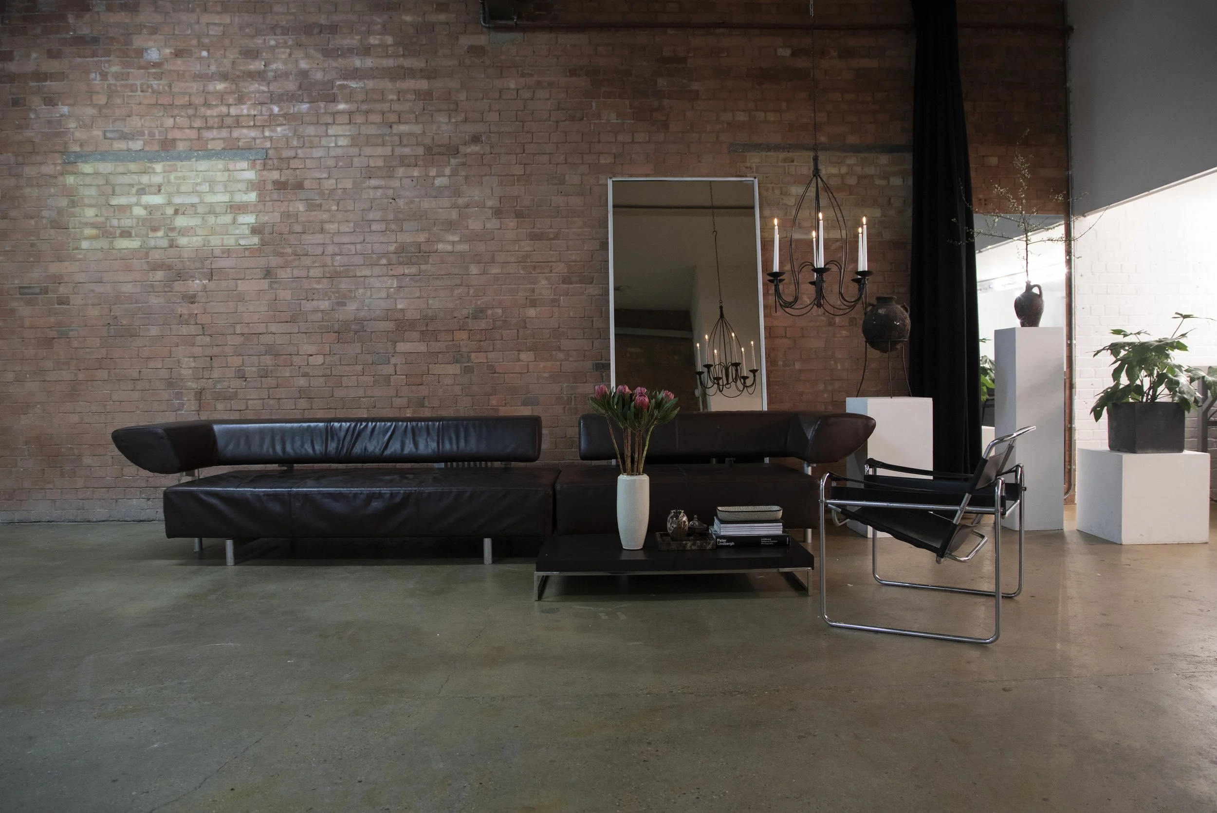 Modern living room with brick wall, black leather sofa, glass coffee table with books and a white vase with pink flowers, black armchair, tall mirror, chandelier, black curtain, large window with natural light, and potted plants.