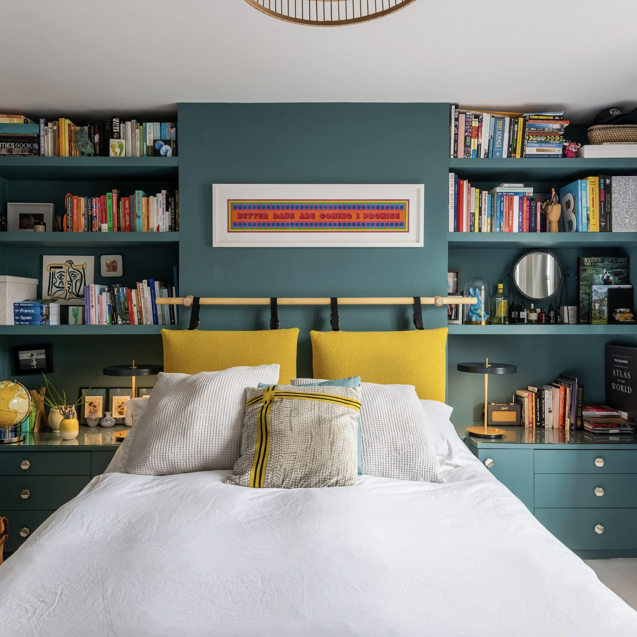 A bedroom with a teal painted wall behind a bed with white bedding and yellow headboard pillows. The wall has built-in bookshelves filled with books, decorations, and framed photos. There are black bedside tables with matching lamps, and a framed col