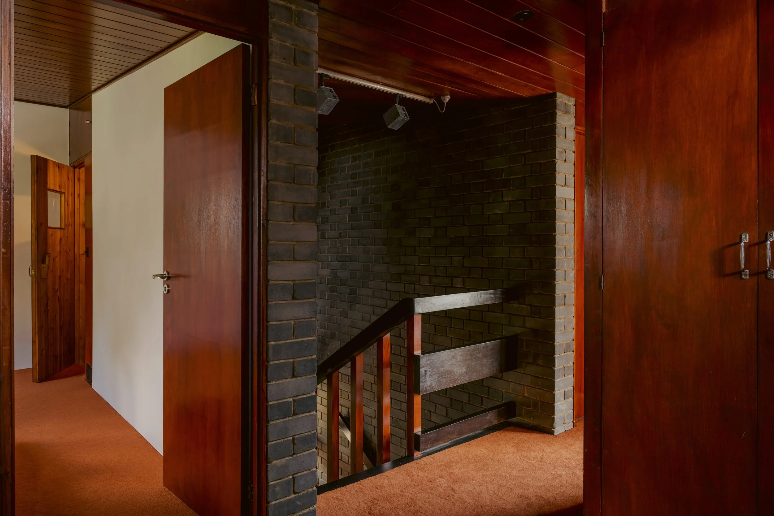 Interior view of a house hallway with wooden doors, brick walls, and a staircase with a wooden handrail. The hallway has orange carpet flooring and wood-paneled ceiling.