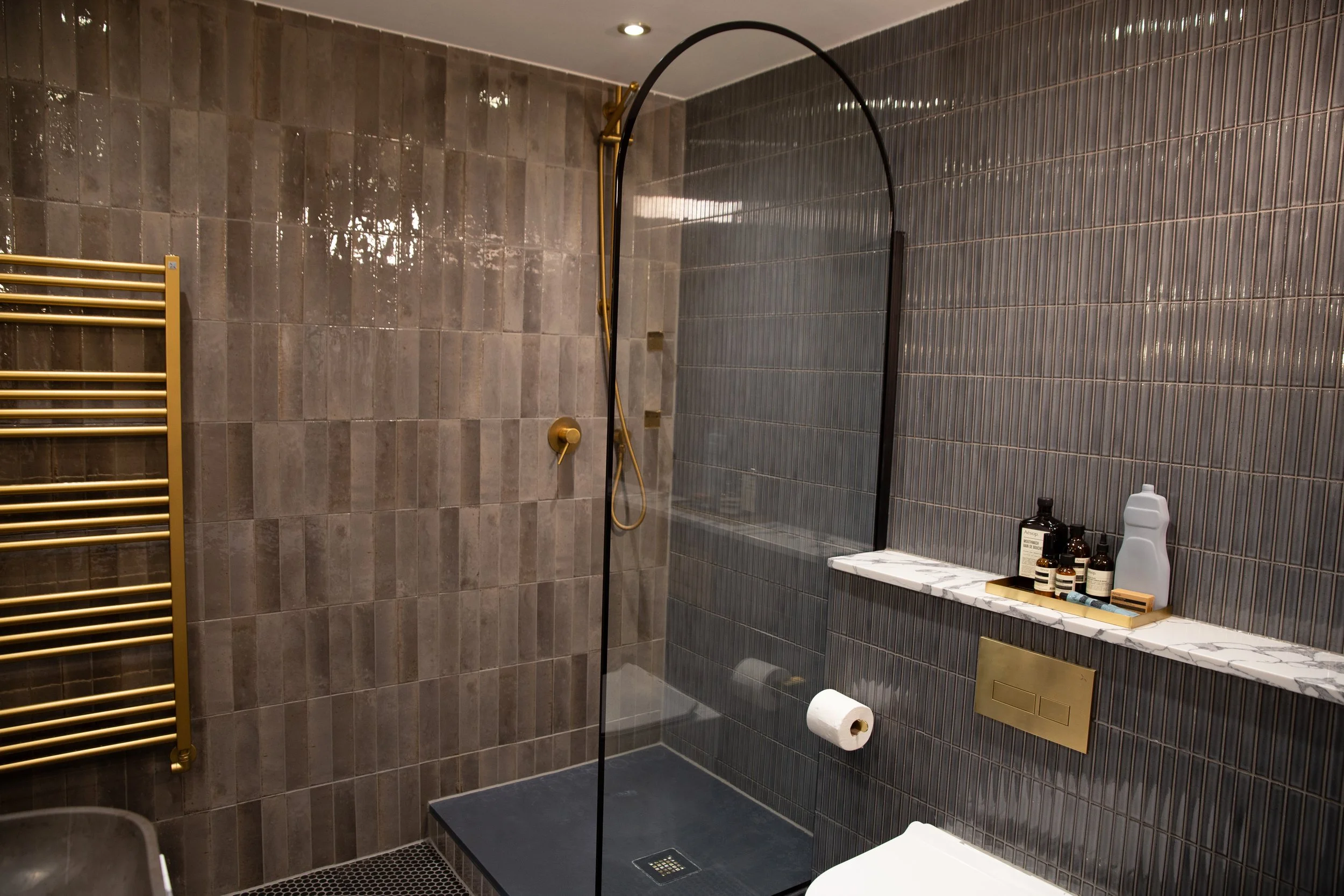 Modern shower area with dark gray tiled walls, a black framed glass door, and brass fixtures, including a wall-mounted showerhead and heating towel rail. A marble shelf holds various toiletries, and a toilet paper roll is mounted on the wall.