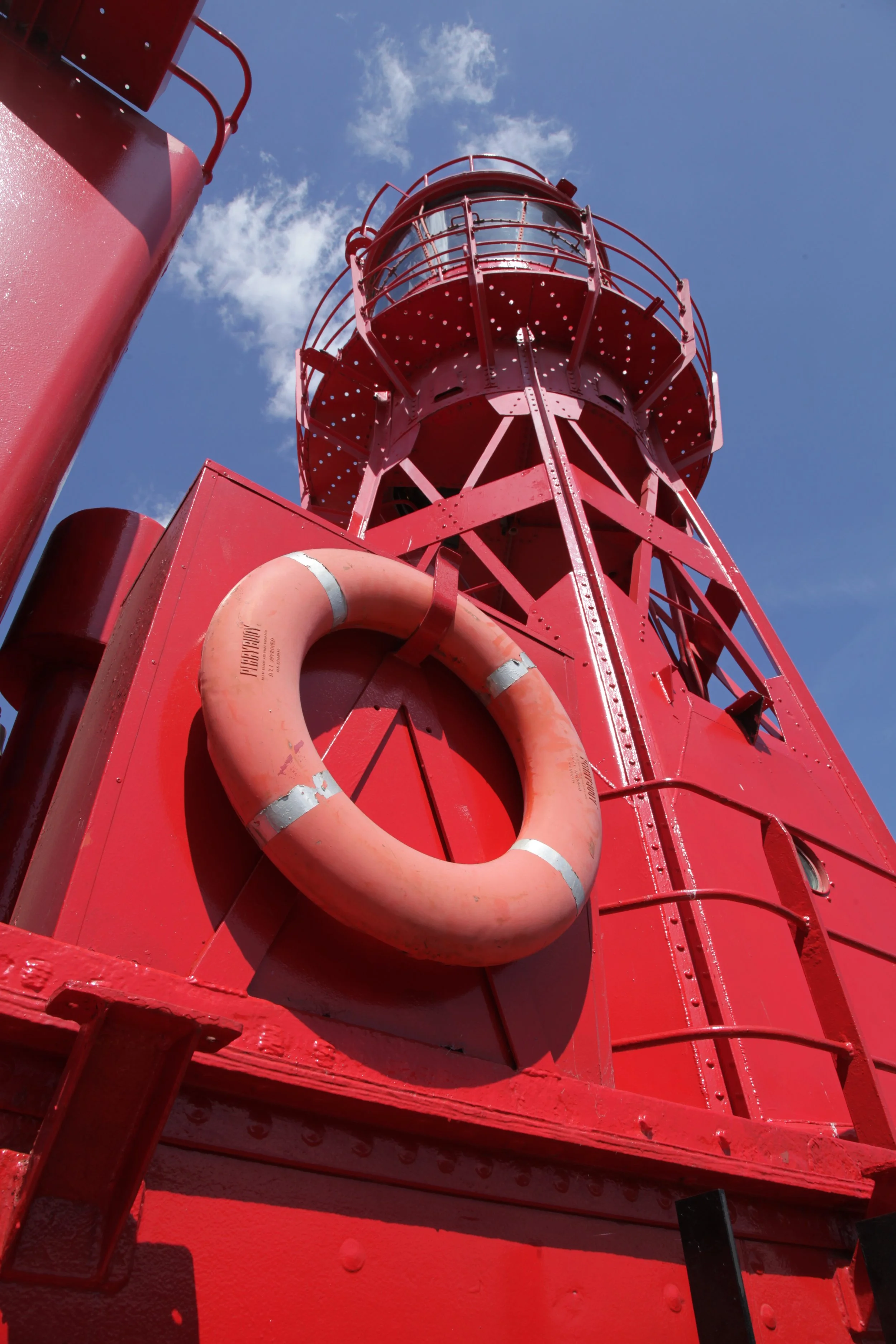 A red lighthouse tower with a life preserver hanging on the side, against a blue sky with white clouds.