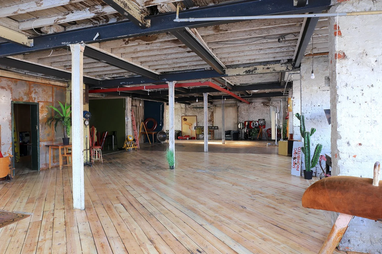 An industrial-style open room with wooden floors, exposed brick and wooden beams, and various furniture and objects including plants, chairs, and equipment.