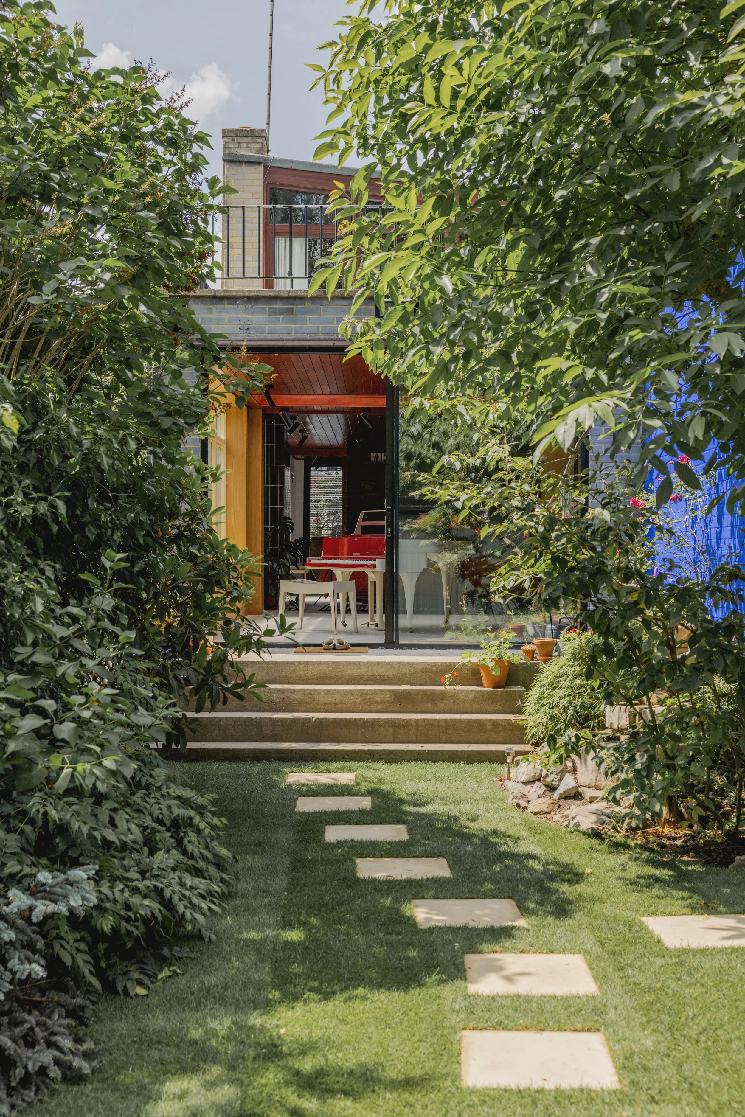 A backyard leading to a house with a glass door, showing a red piano, chairs, and potted plants inside, with a stepping stone path on a green lawn and dense bushes on each side.