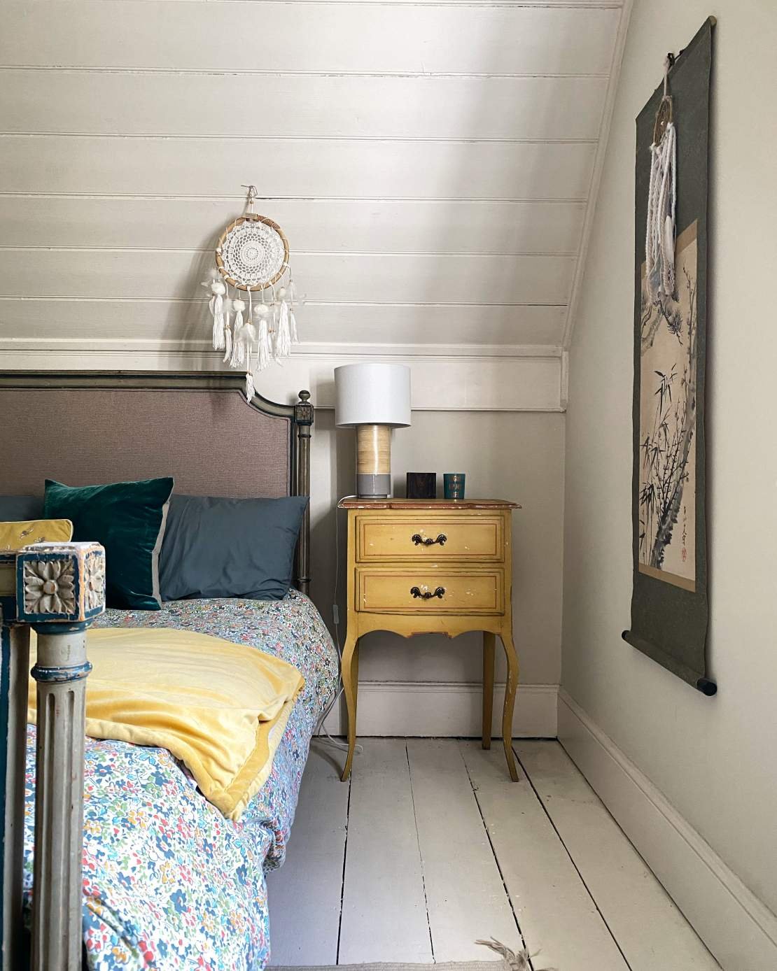 A bedroom corner featuring a bed with colorful bedding, a yellow nightstand with black handles, a white lamp, a dreamcatcher hanging on the wall, and a hanging scroll with traditional Asian art on the wall.