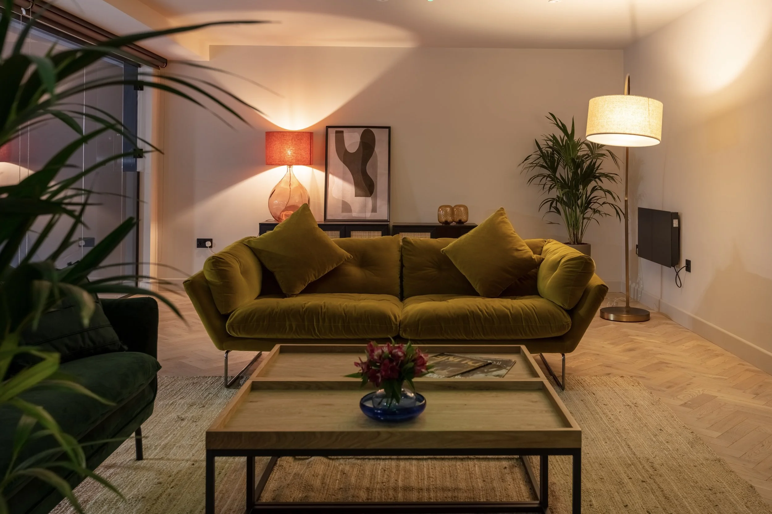 Cozy living room with a mustard yellow sofa, a wooden coffee table with a flower vase, a green armchair, potted plants, lamps, and wall art.
