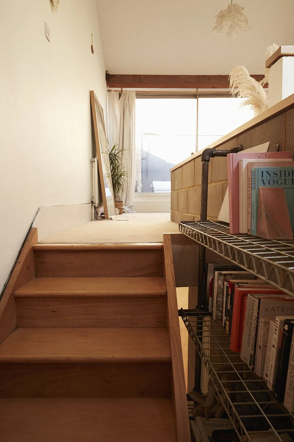 Interior of a home with wooden stairs leading to a bright room with large window, curtain, mirror, potted plant, bookshelf, and decorative plants.
