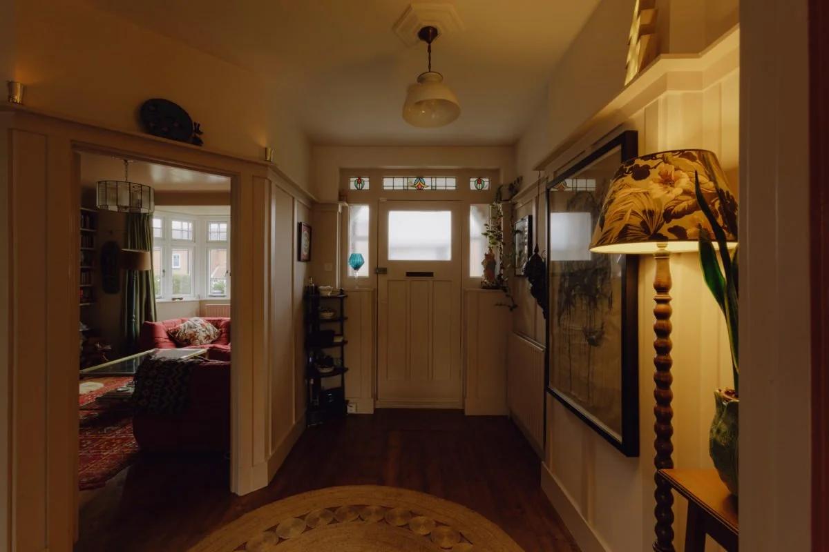Interior view of a hallway leading to a front door with stained glass panels, with a living room visible through an open doorway on the left, decorated with cushions, curtains, and a hanging lamp.