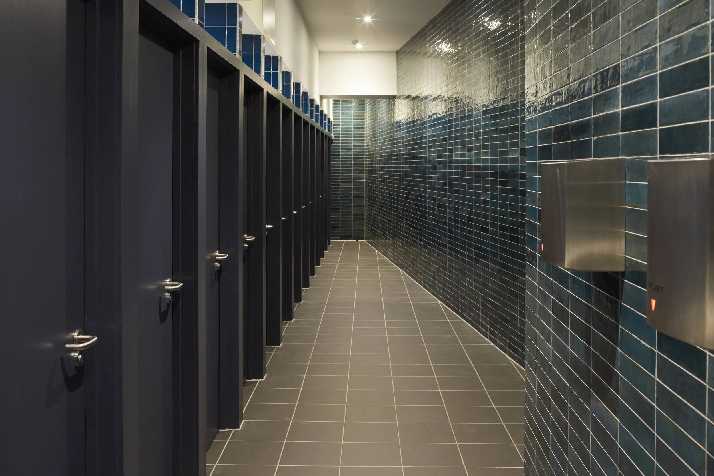 A row of dark bathroom stalls along a tiled wall and floor in a modern public restroom.