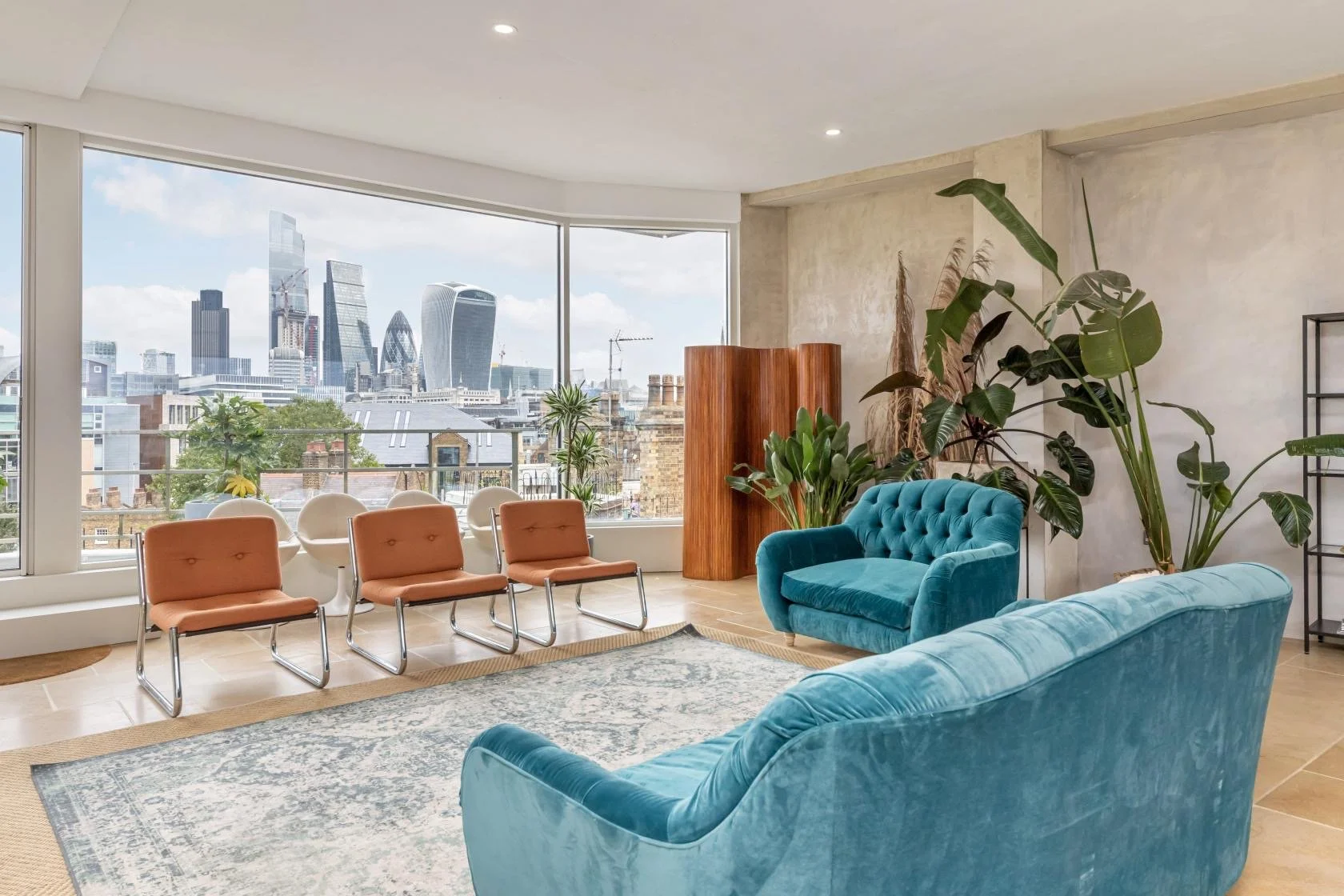 Modern living room with large window showcasing a city skyline, green plants, blue velvet sofas, a patterned area rug, and brown chairs.