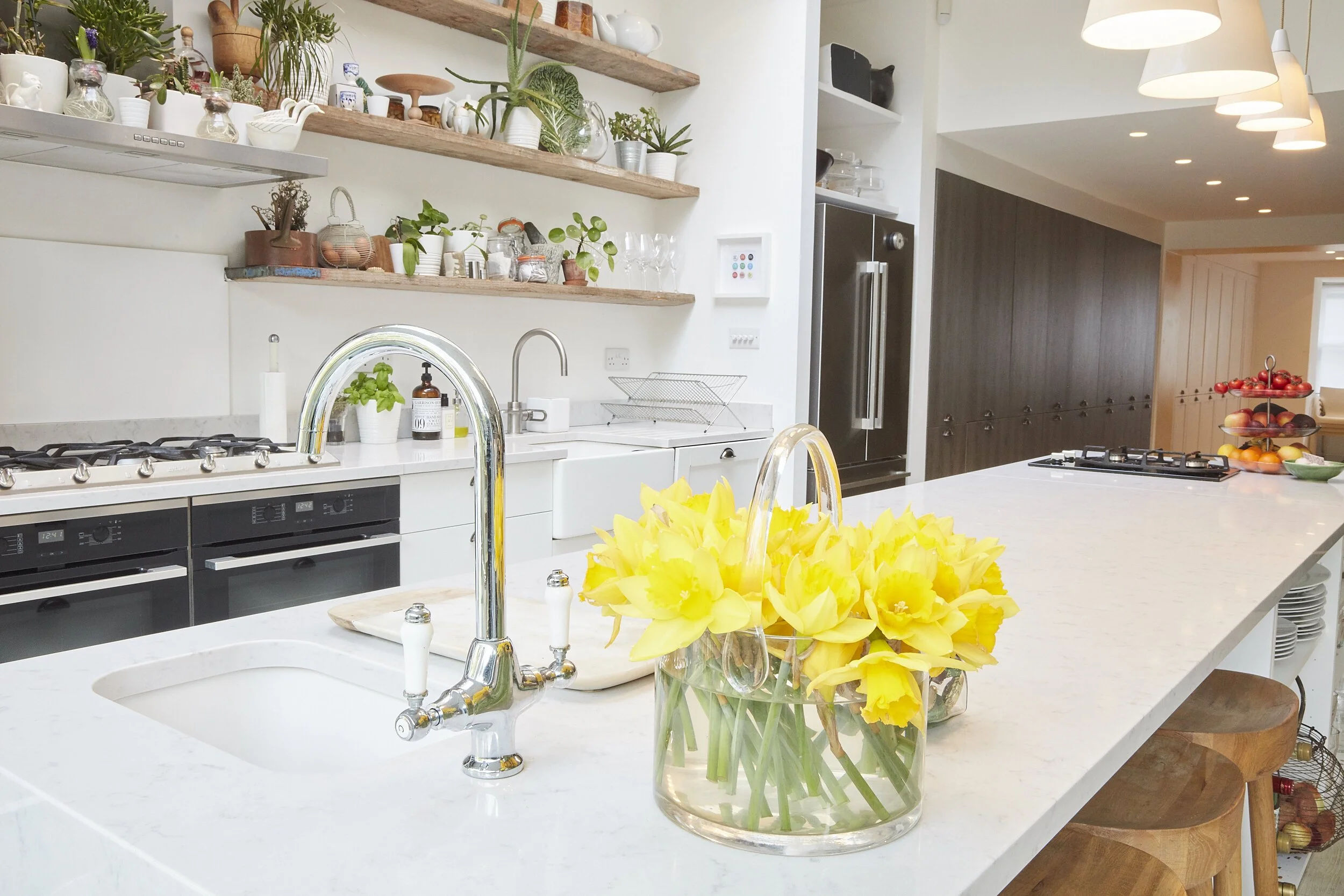 A modern kitchen with white countertops, open wooden shelves with plants and jars, stainless steel appliances, a bowl of yellow daffodils, and a breakfast bar with wooden stools.