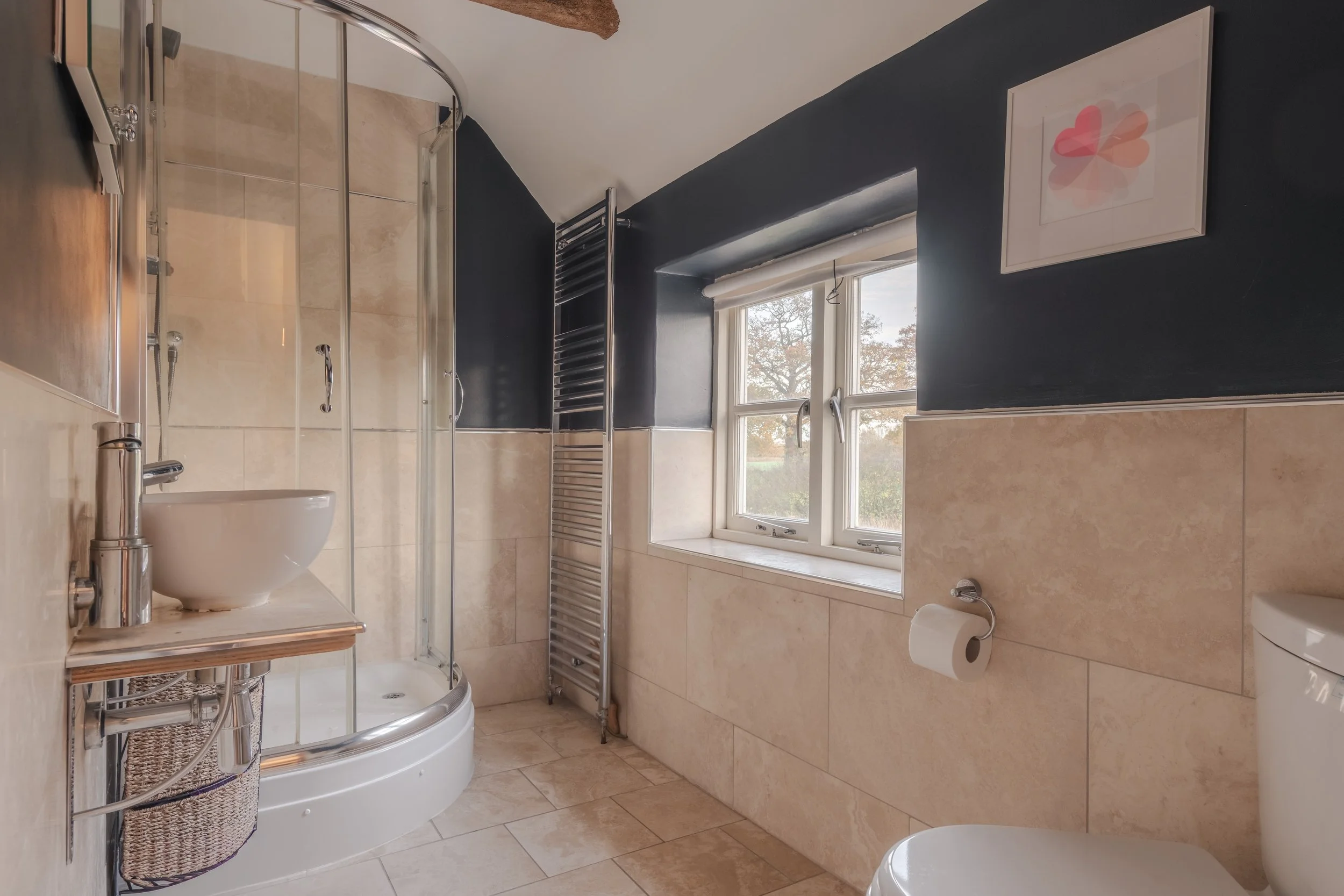 Modern bathroom with a corner shower enclosure, a small white sink, a towel radiator, a window with a roman shade, a framed art piece on a dark blue wall, and a toilet.