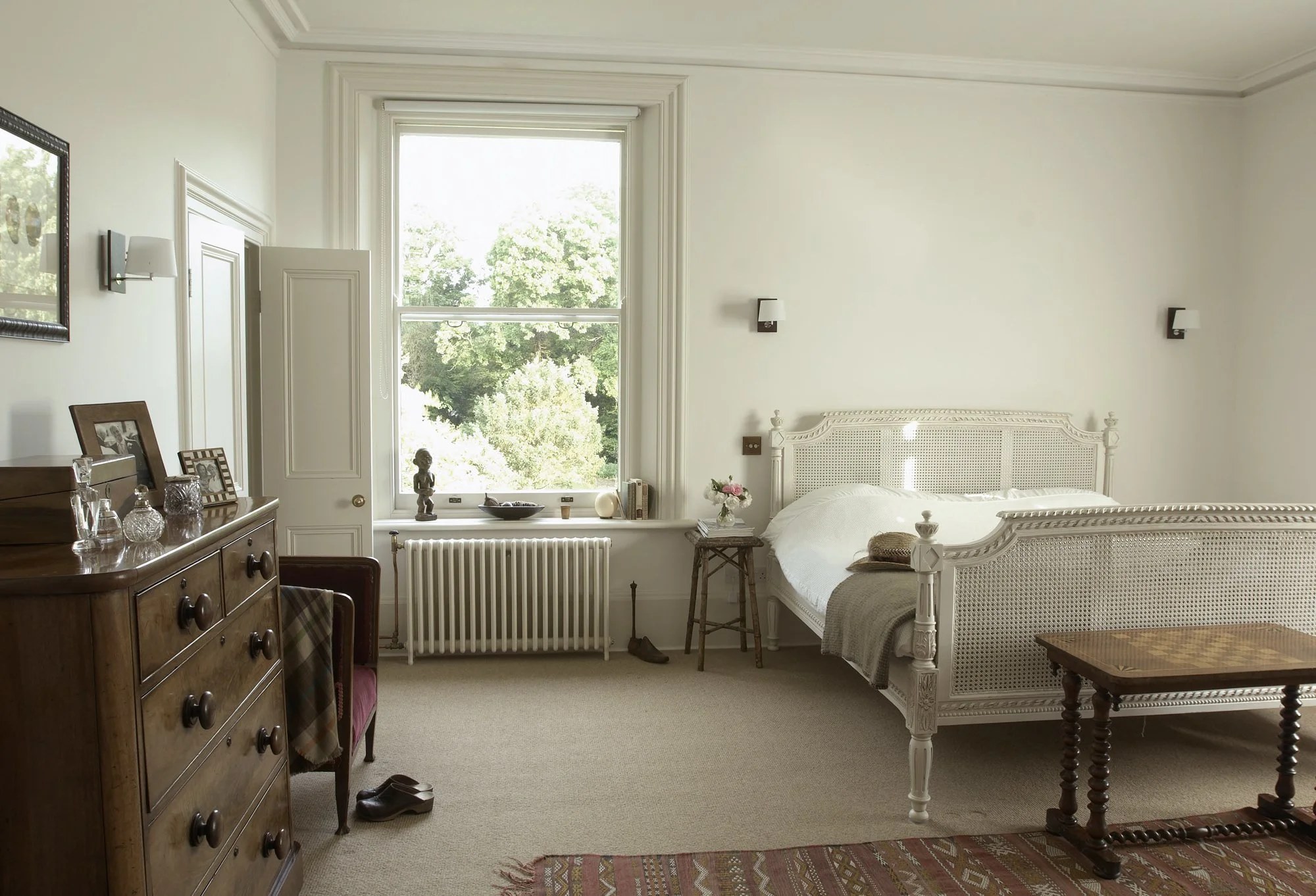 A bedroom with white walls, a large window showing green trees outside, a white bed with cane detail, a wooden chest of drawers with framed photos and glassware, a small side table with a vase of flowers, a wooden table at the foot of the bed, and a 