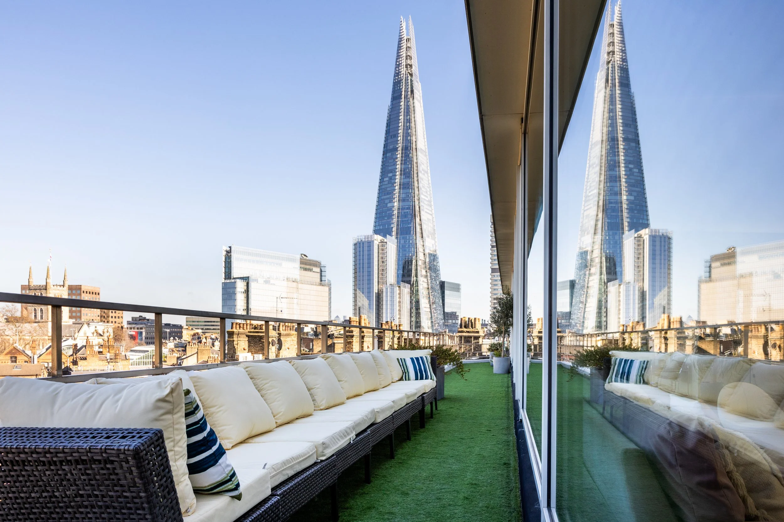 Rooftop balcony with white cushioned seating along the edge, potted plants, and cityscape view of modern glass skyscrapers including The Shard in London.