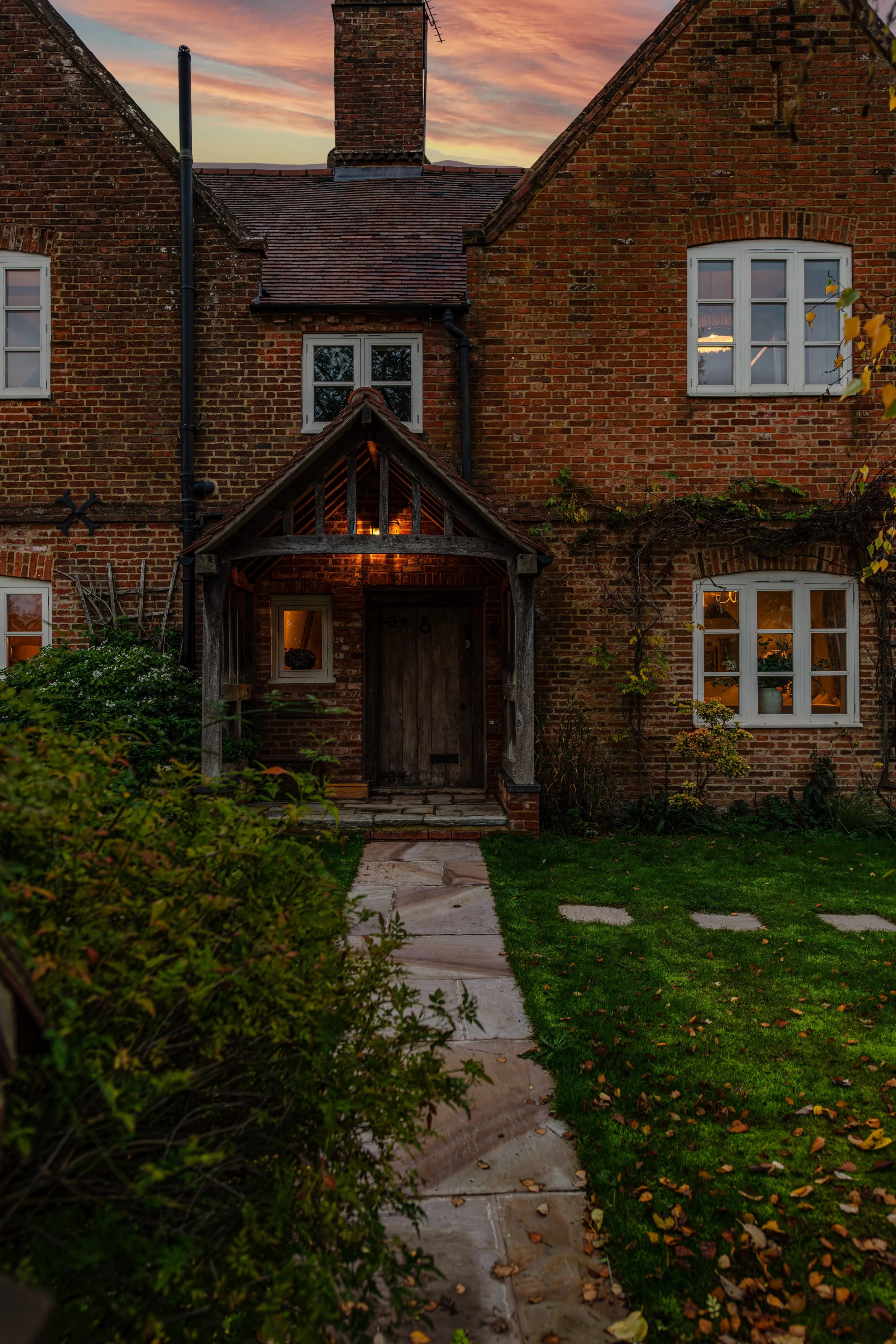 A brick house with a wooden front door, a small porch, and white-framed windows. The doorway is illuminated with warm light, and there are illuminated windows in the house. The sky is at sunset or dusk, with shades of pink, purple, and orange. There 