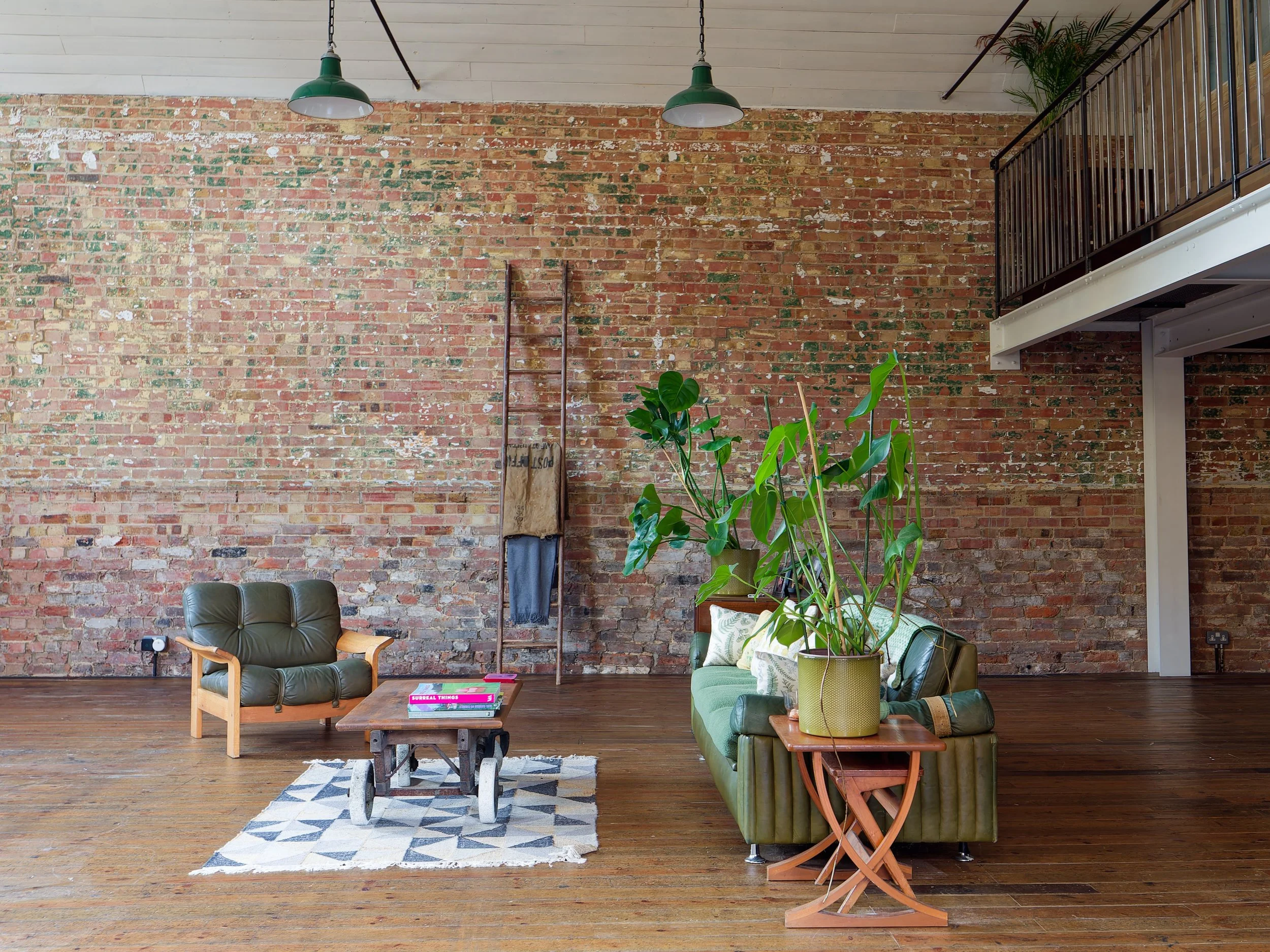 Living room with a brick wall, a green armchair, a green sofa, a wooden coffee table, a large potted plant, and industrial-style ceiling lights.