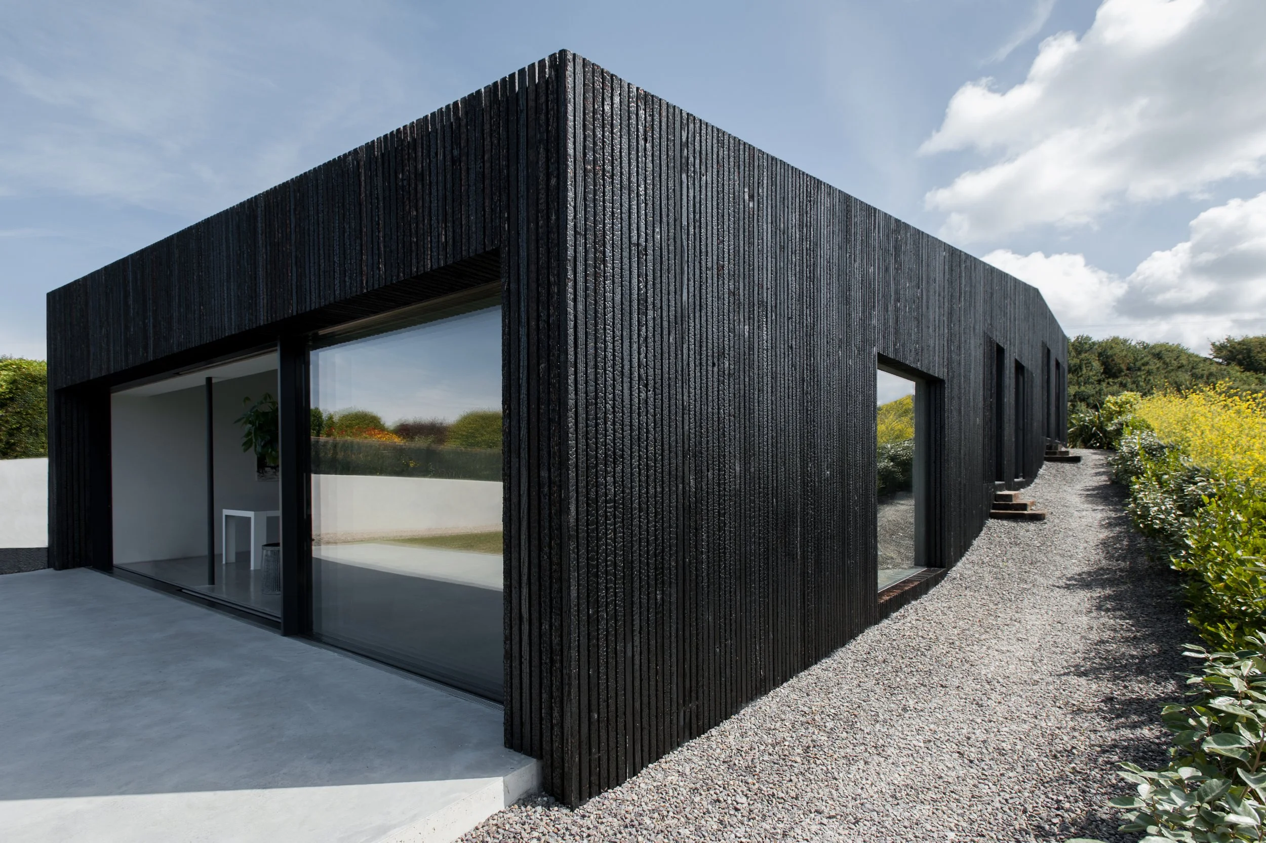 Modern black house with large glass sliding doors, gravel pathway on right side, and outdoor patio with minimalistic furniture, surrounded by greenery and cloudy sky.