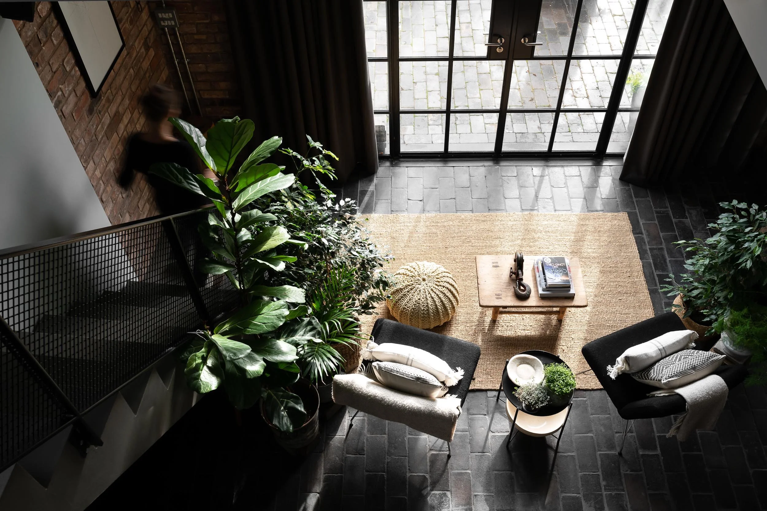 Top-down view of a modern living room with black brick flooring, a beige rug, two black armchairs with white scarves and pillows, a small round table with green plants, a wooden coffee table with books and a vintage phone, large green plants in the c