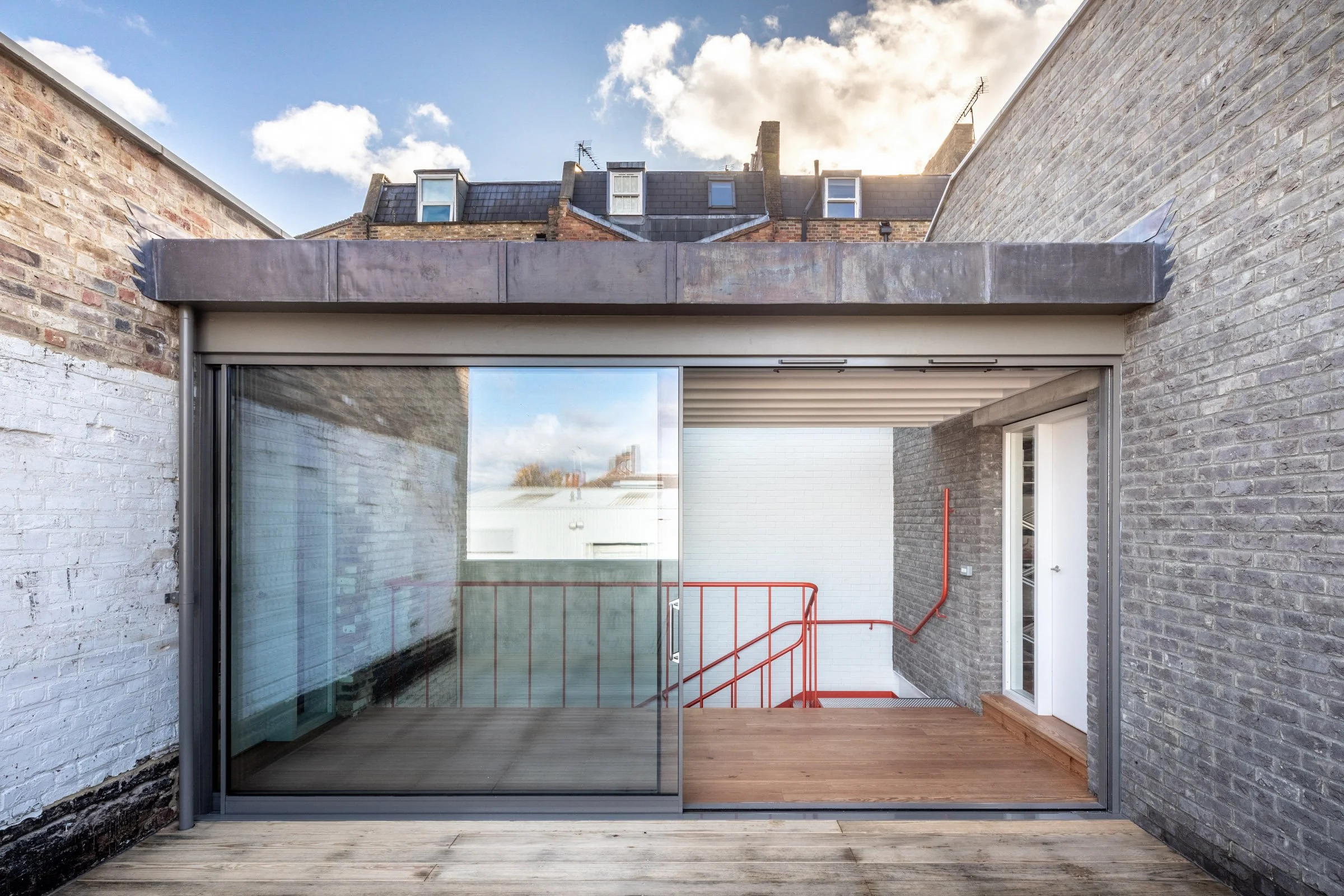 Urban building with a glass sliding door leading to an outdoor wooden deck, overlooking a staircase with red railings and neighboring rooftops under a partly cloudy sky.
