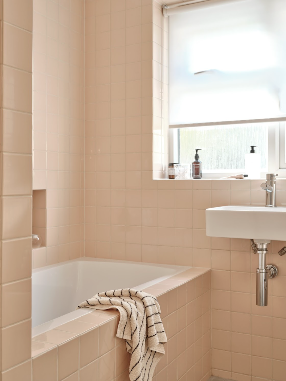 A bathroom with peach-colored tiled walls, a white bathtub with a striped towel hanging over its edge, a small white sink, and a window with a frosted window covering, with soap dispensers and toiletries on the window sill.