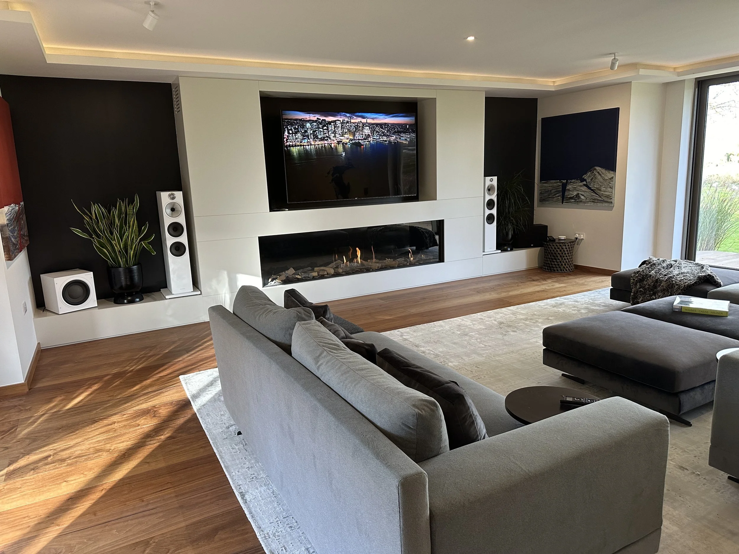 Modern living room with a gray sofa, black ottoman, large area rug, built-in fireplace, flat-screen TV, and sliding glass doors showing a green outdoor area.