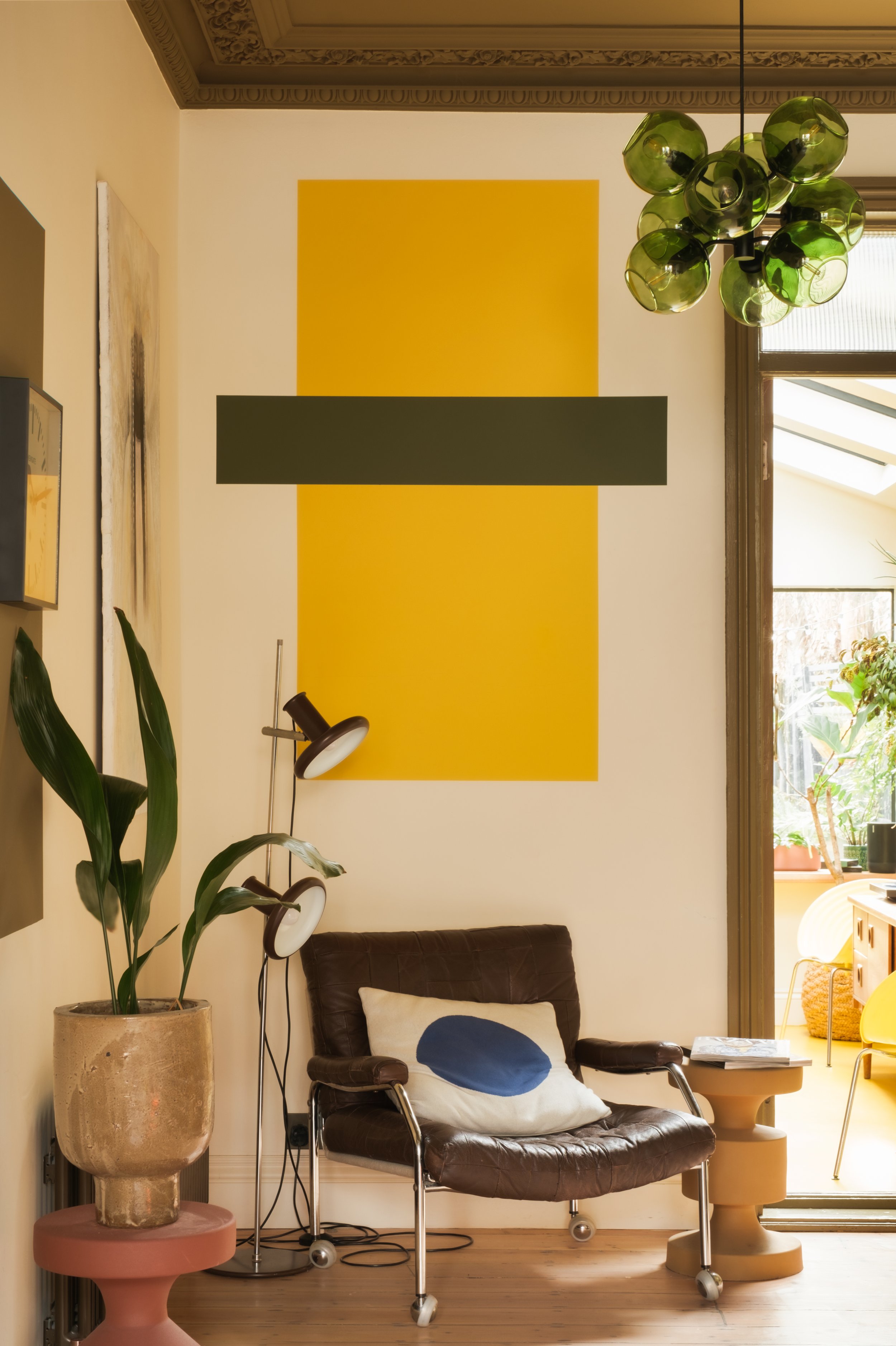 Living room with a brown leather armchair, a beige pillow with a blue circle, a side table with a book, a potted plant, and a floor lamp. There is a large yellow and gray abstract wall art, a green glass chandelier, and a sliding glass door leading t