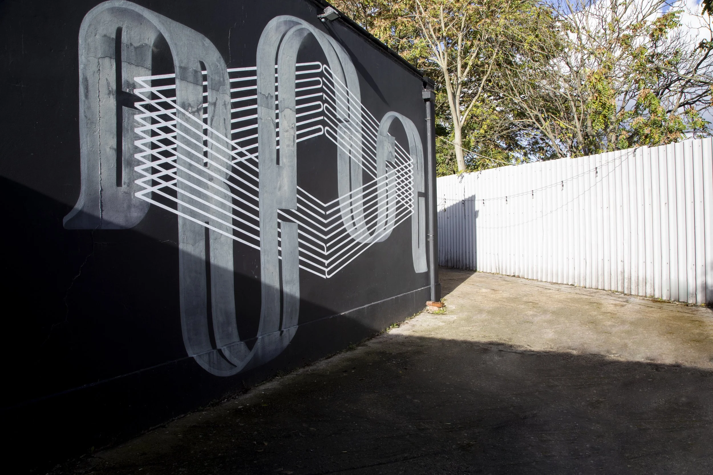 Street art mural painted on a black wall, featuring large, three-dimensional, stylized gray letters with white geometric lines connecting them.