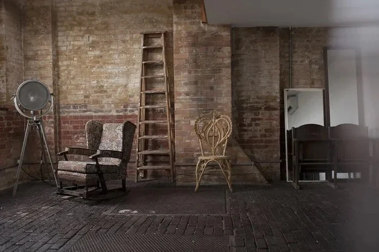 Interior of a room with a brick wall, an old armchair, a vintage spotlight, a wooden ladder, a metal chair, and a large mirror.